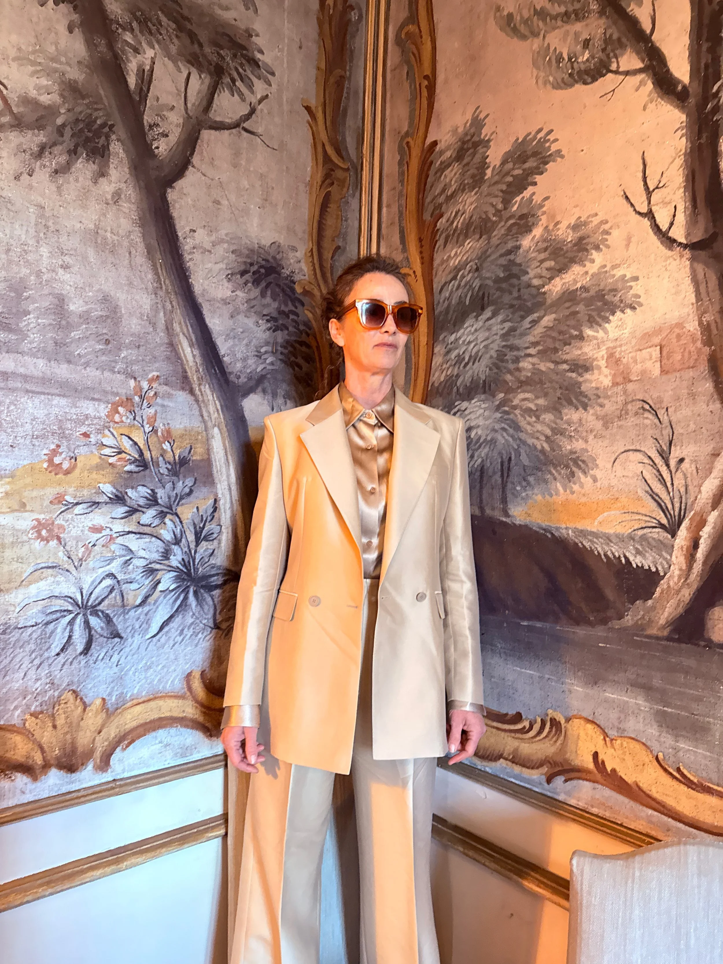 Woman in a light tan suit and sunglasses standing in front of an ornate, large wall mural of trees and foliage painted in earthy tones.
