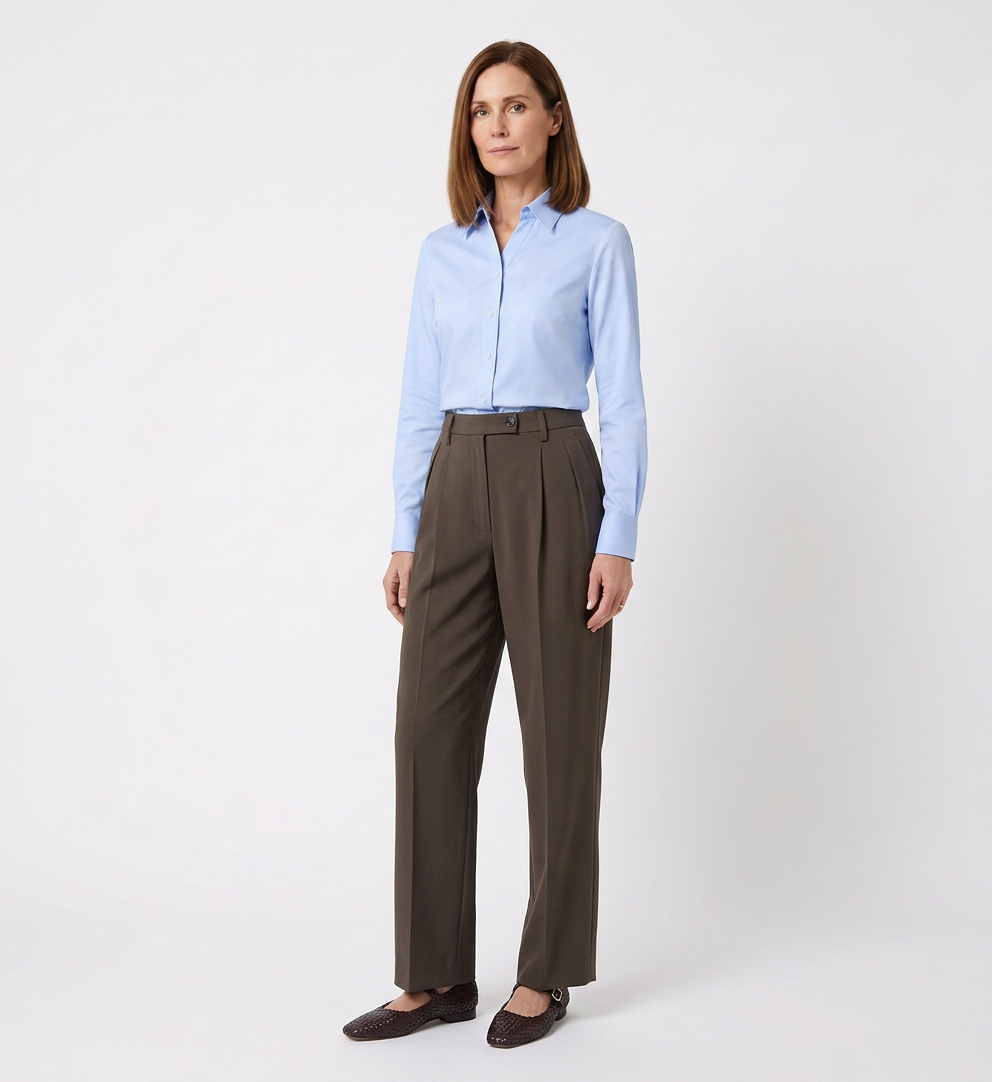 A woman standing against a plain white background, wearing a light blue button-up shirt, dark brown high-waisted trousers, and dark brown shoes.