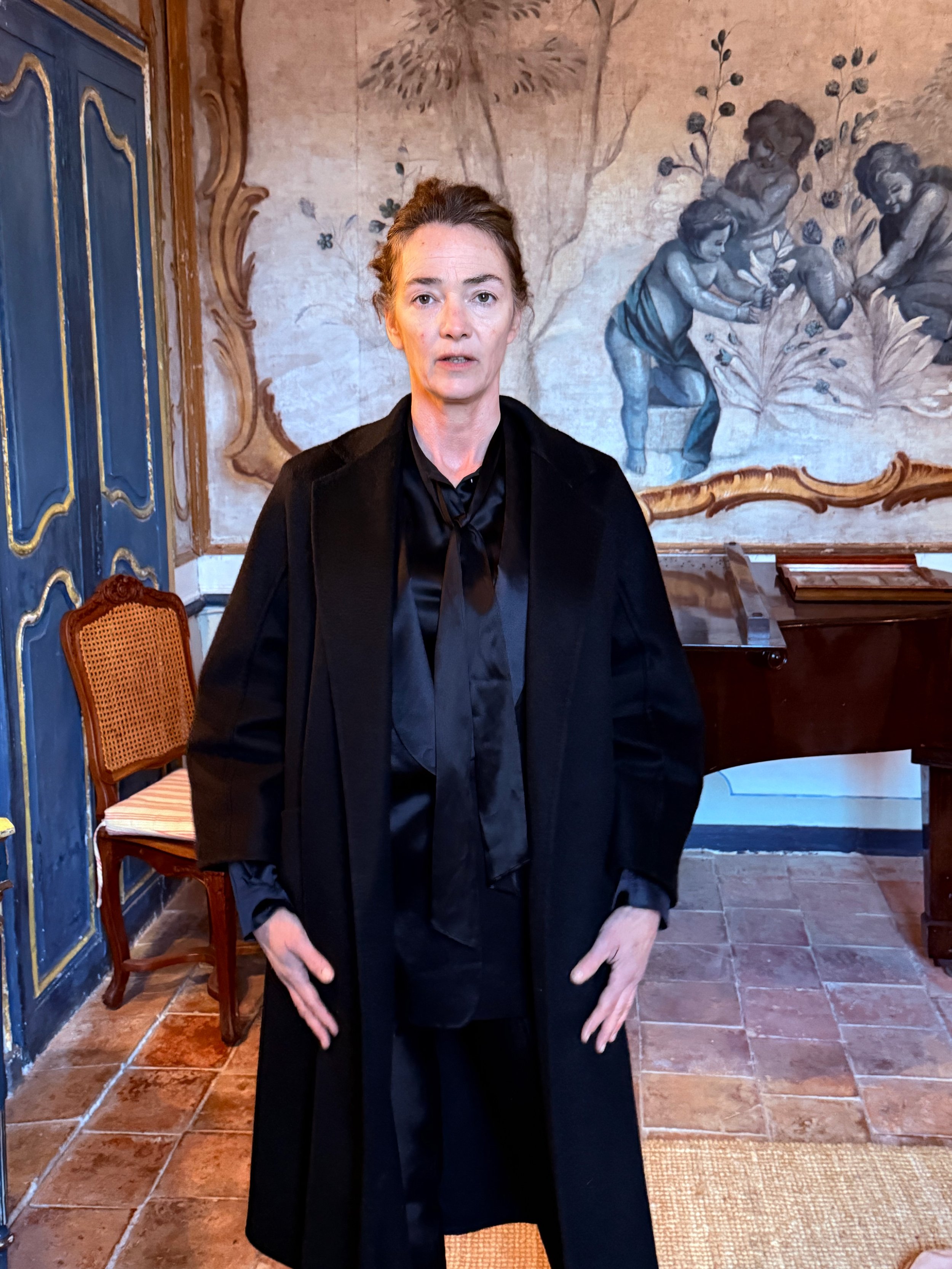 A woman with brown hair styled in an updo, wearing a black jacket over a black shirt, standing in a room with a painted wall depicting children and plants, next to a blue wooden door with gold trim and a wooden chair with a woven backrest.
