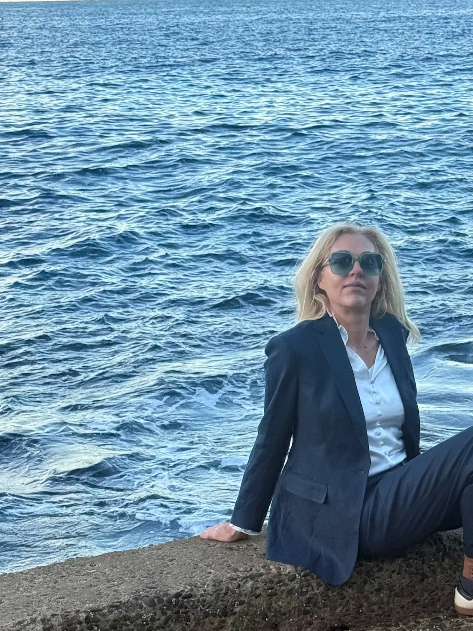 A woman with blonde hair wearing sunglasses, a white shirt, and a dark blazer is sitting on a rocky beach near the ocean, with her right arm resting on the rocks and her left arm bent at the elbow. The water behind her is blue with gentle waves.