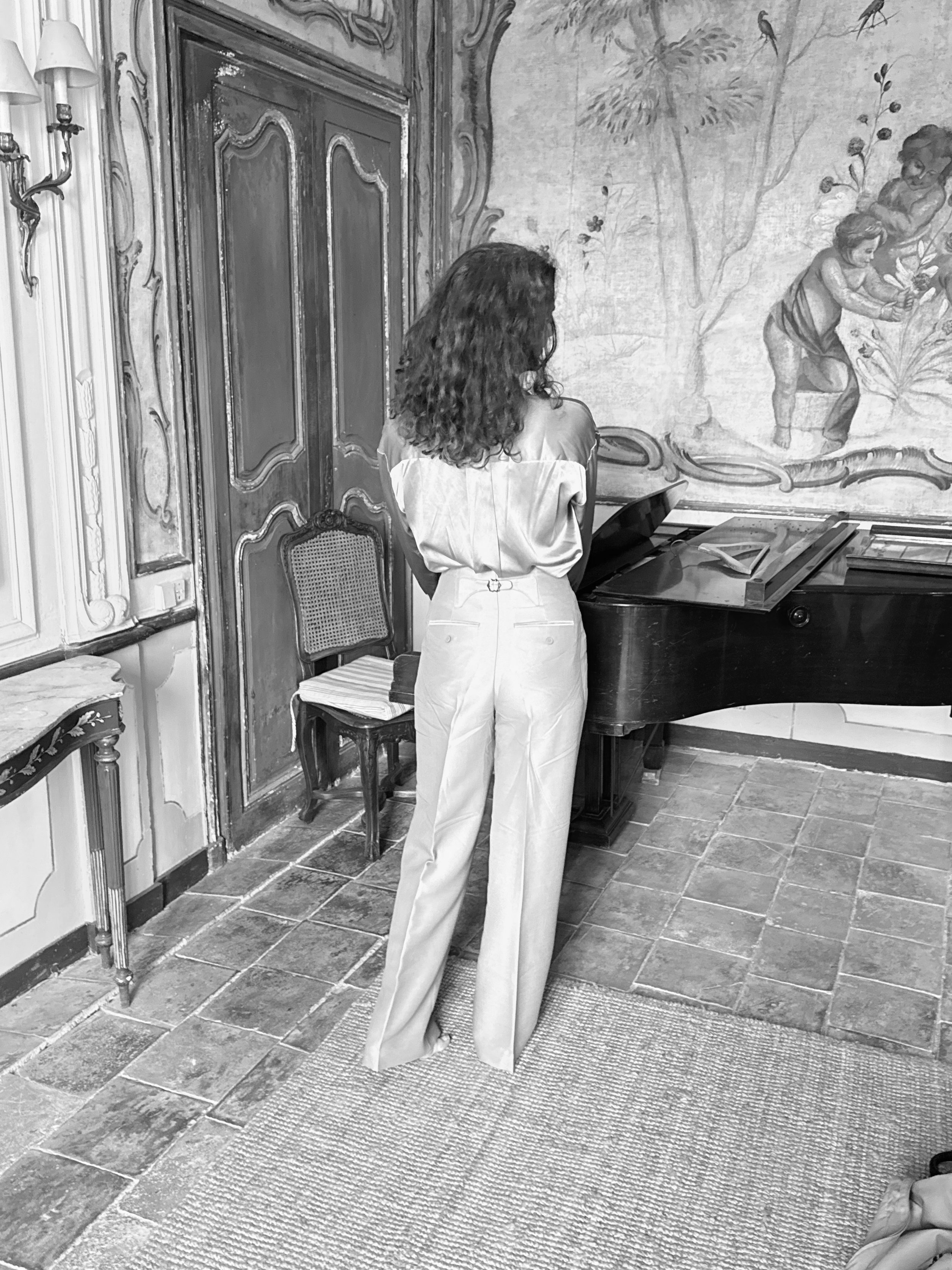 A woman with curly hair wearing light-colored clothing stands in a room with ornate wall decor, a mural painting of children playing in a garden, and a grand piano. She is facing away from the camera, looking toward the mural.