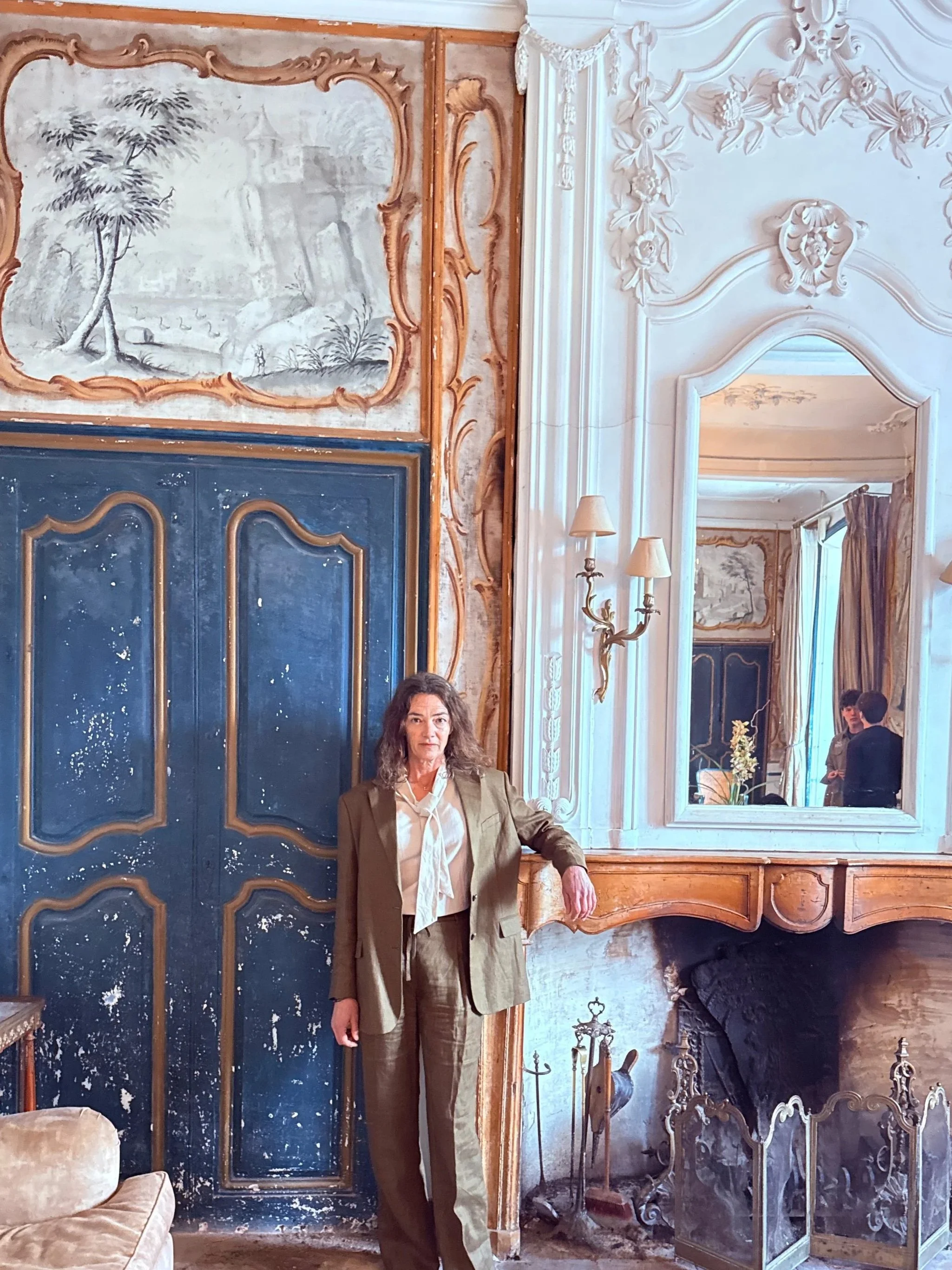 A woman with long curly hair wearing a beige suit and white blouse standing in a room with ornate interiors. The room features decorative molding, a mirror, a large painting or mural with a landscape scene, and vintage furniture including a velvet armchair, a small table, and candlesticks.
