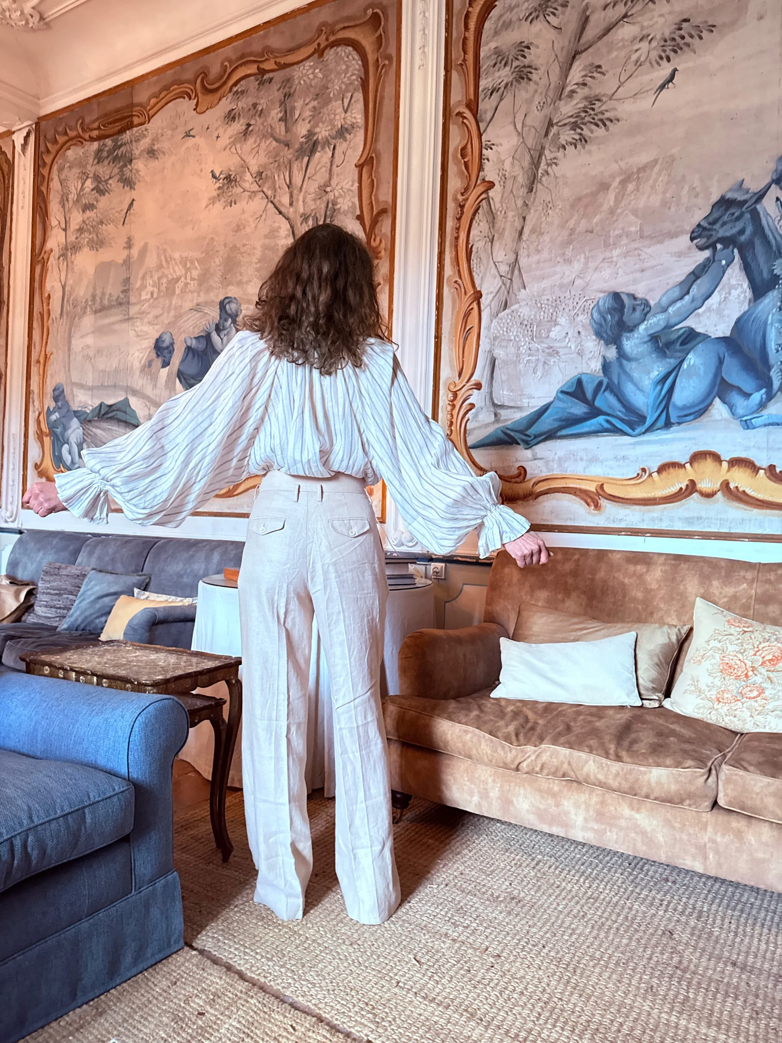 A woman with curly hair, wearing a light-colored, striped blouse and white pants, stands in a living room with her back to the camera, facing a large wall-sized mural. The mural depicts a classical scene with people, trees, and a horse, framed with ornate woodwork. The room has various sofas, a wooden table, and cushions on the floor, with a beige carpet covering the floor.