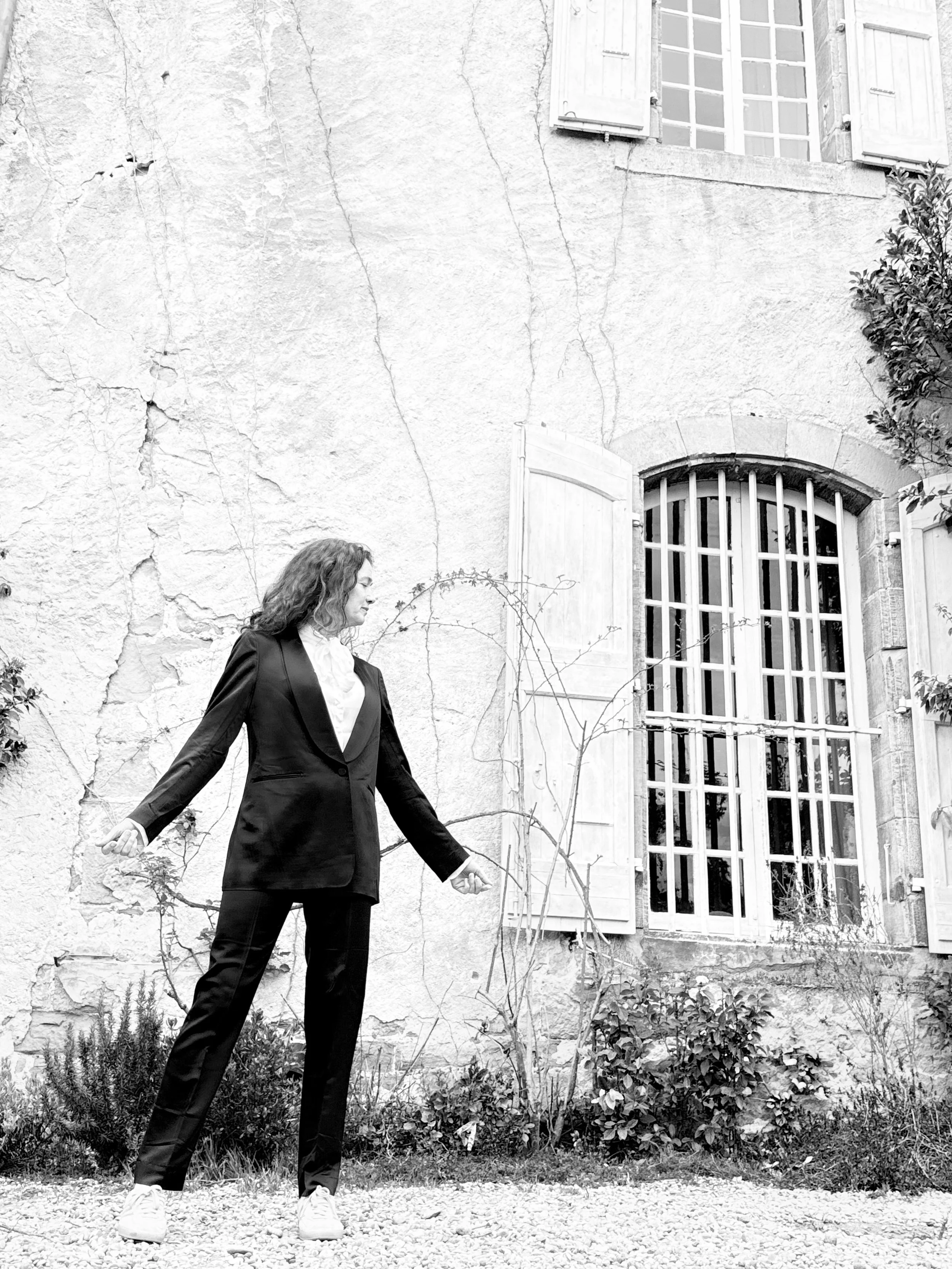 A woman in a suit and sneakers is walking outside on a gravel path along a building with a textured wall, a large window with shutters, and some plants.