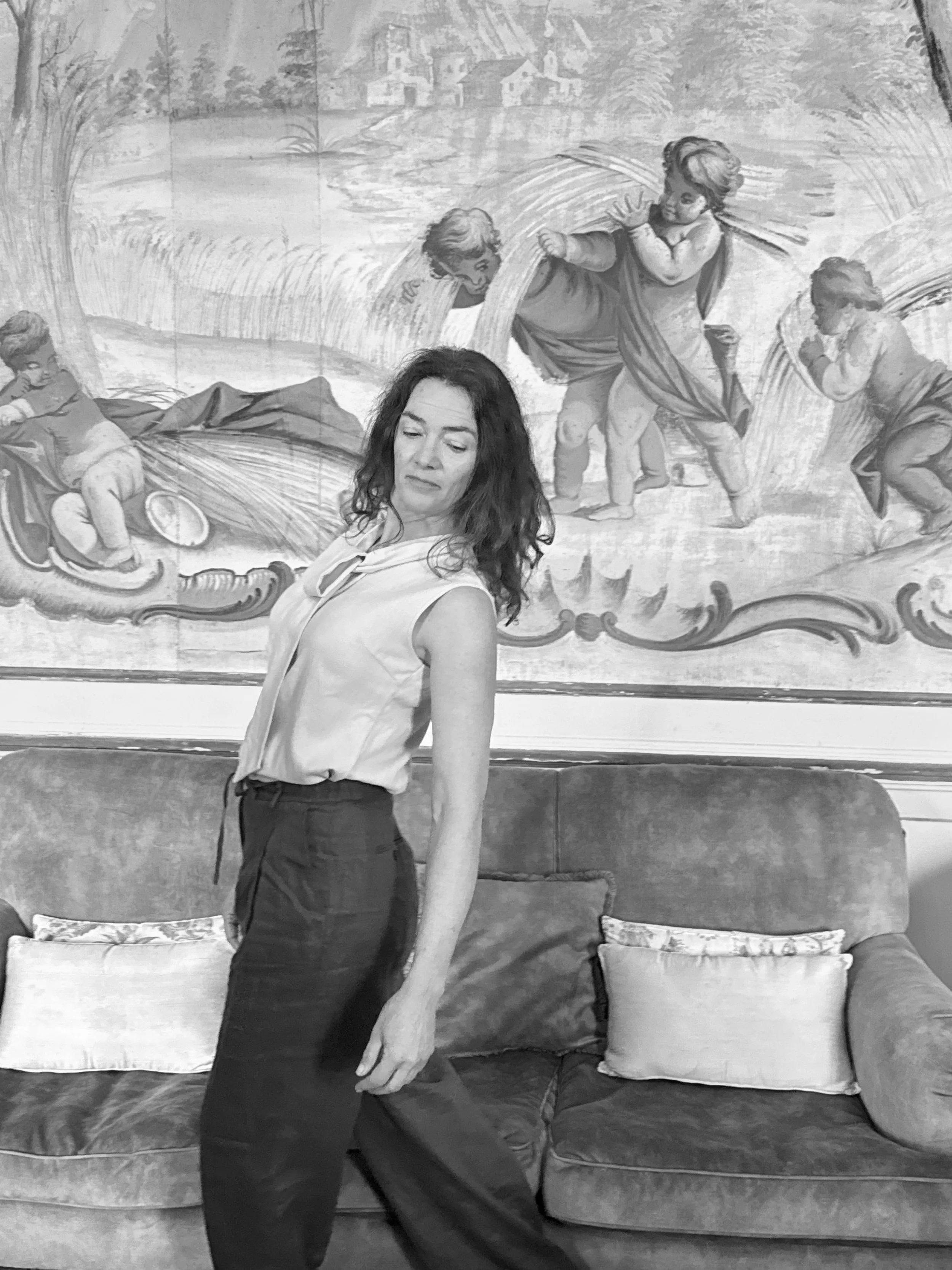 A woman with long, curly hair standing in front of a large painting depicting children playing by a river.