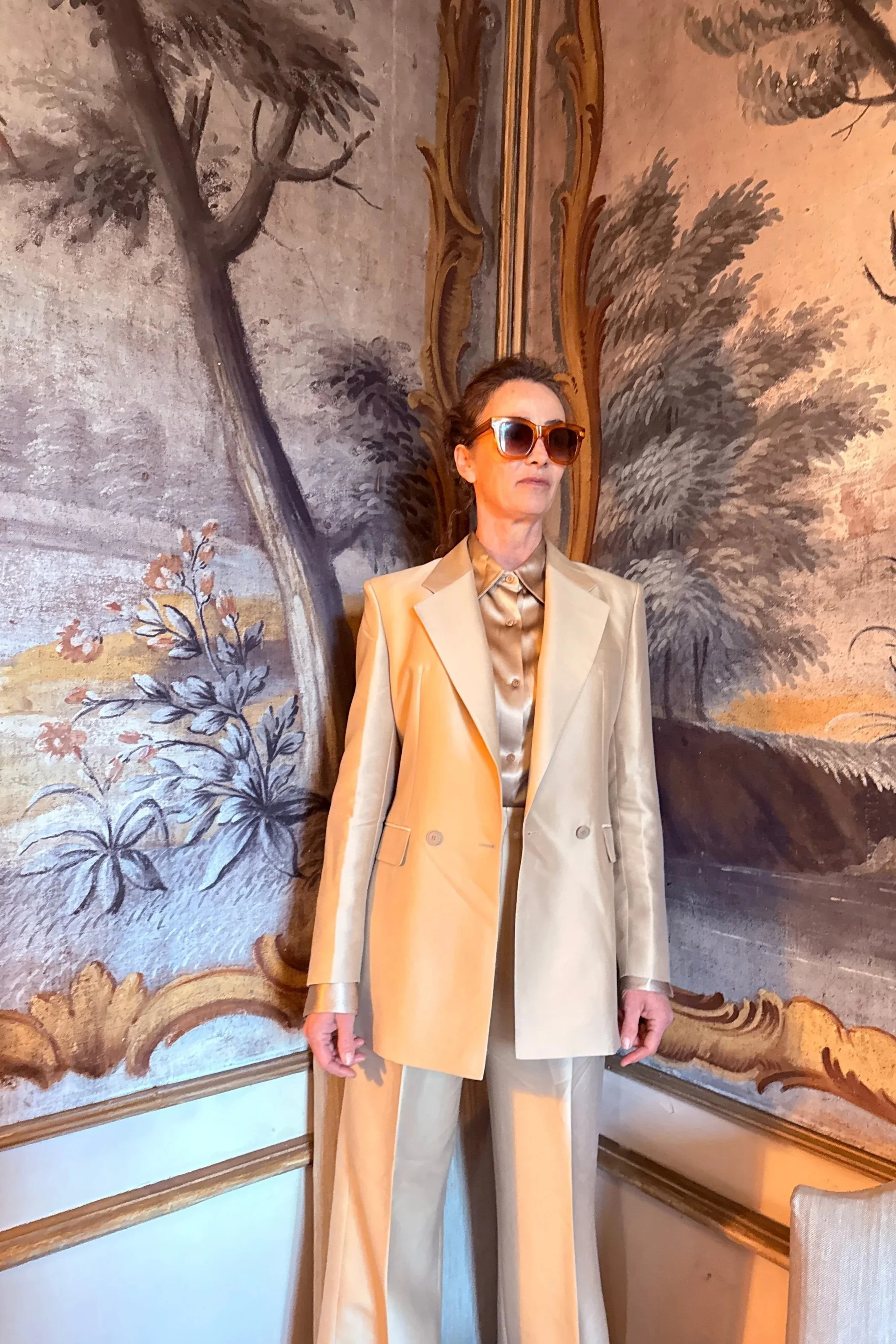 Person wearing large sunglasses, a beige suit jacket over a satin shirt, and beige trousers, standing in front of a decorative, painted wall with trees and floral motifs.