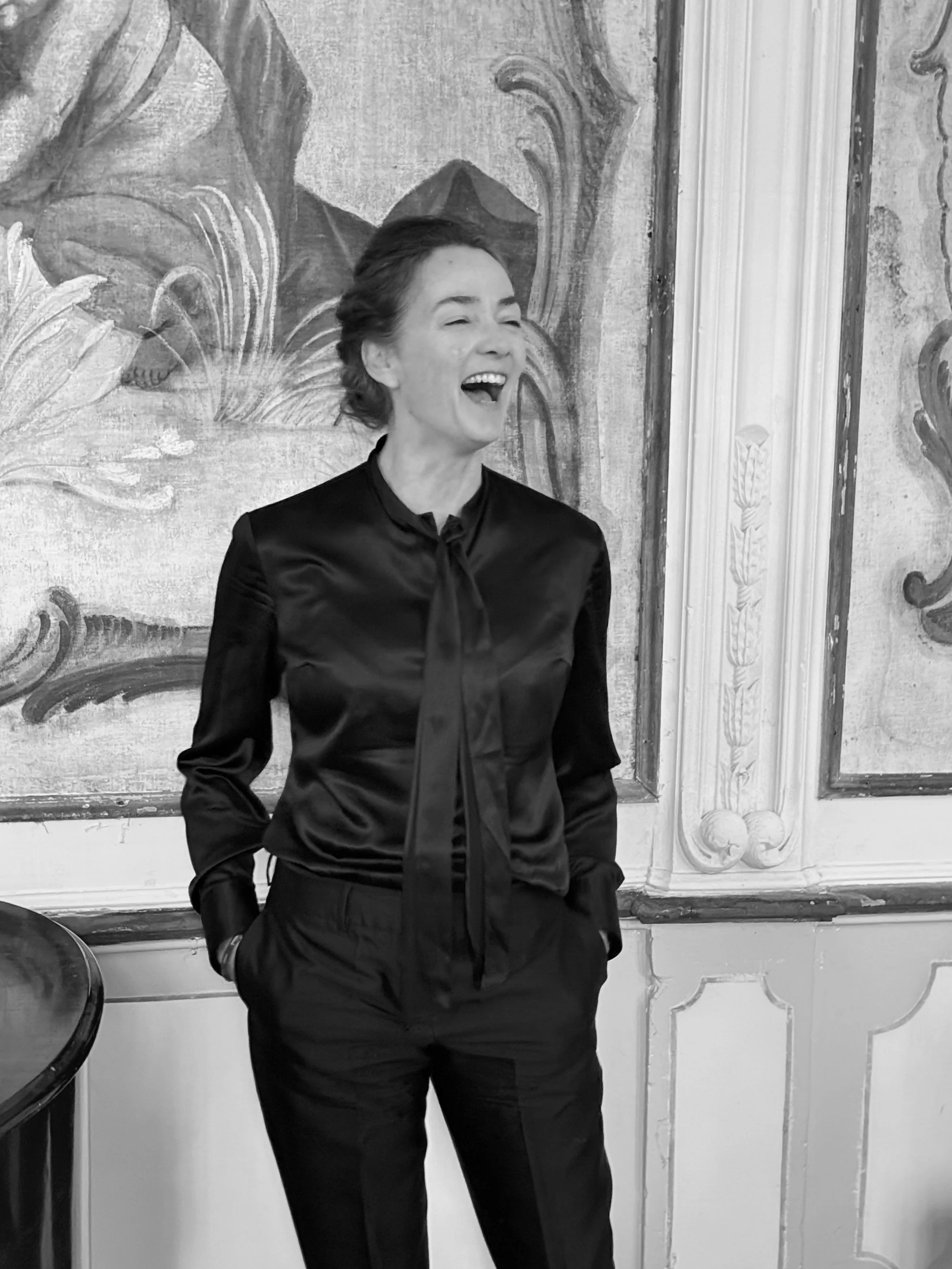 Black and white photo of a woman laughing, wearing a dark satin blouse with a bow tie and dark pants, standing in a room with ornate wall decorations.