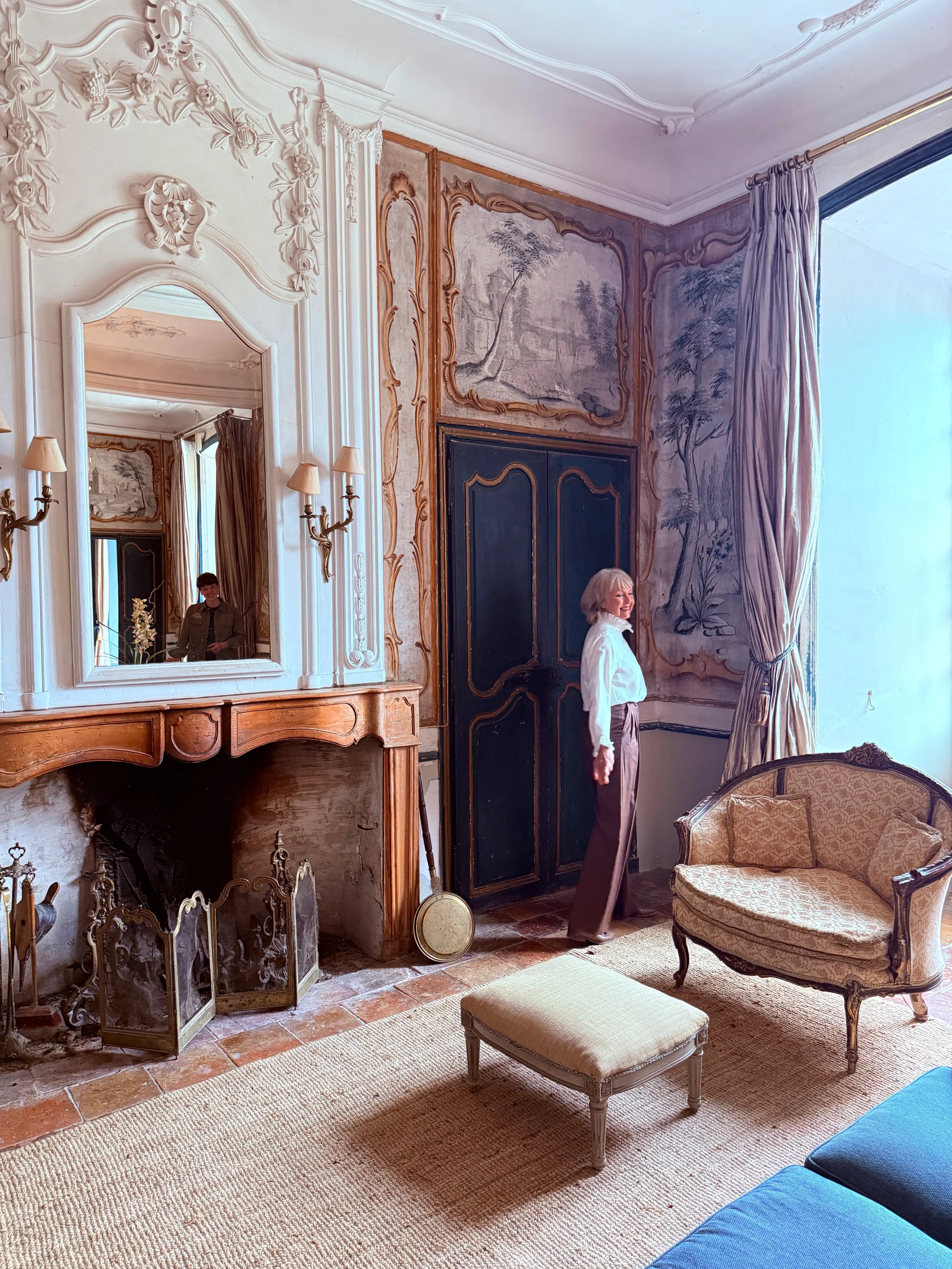 Elegant room with a fireplace, a large mirror, antique furniture including a sofa, armchair, small table, and a woman standing near draped window with large curtains, detailed wall art, and a rug covering the floor.