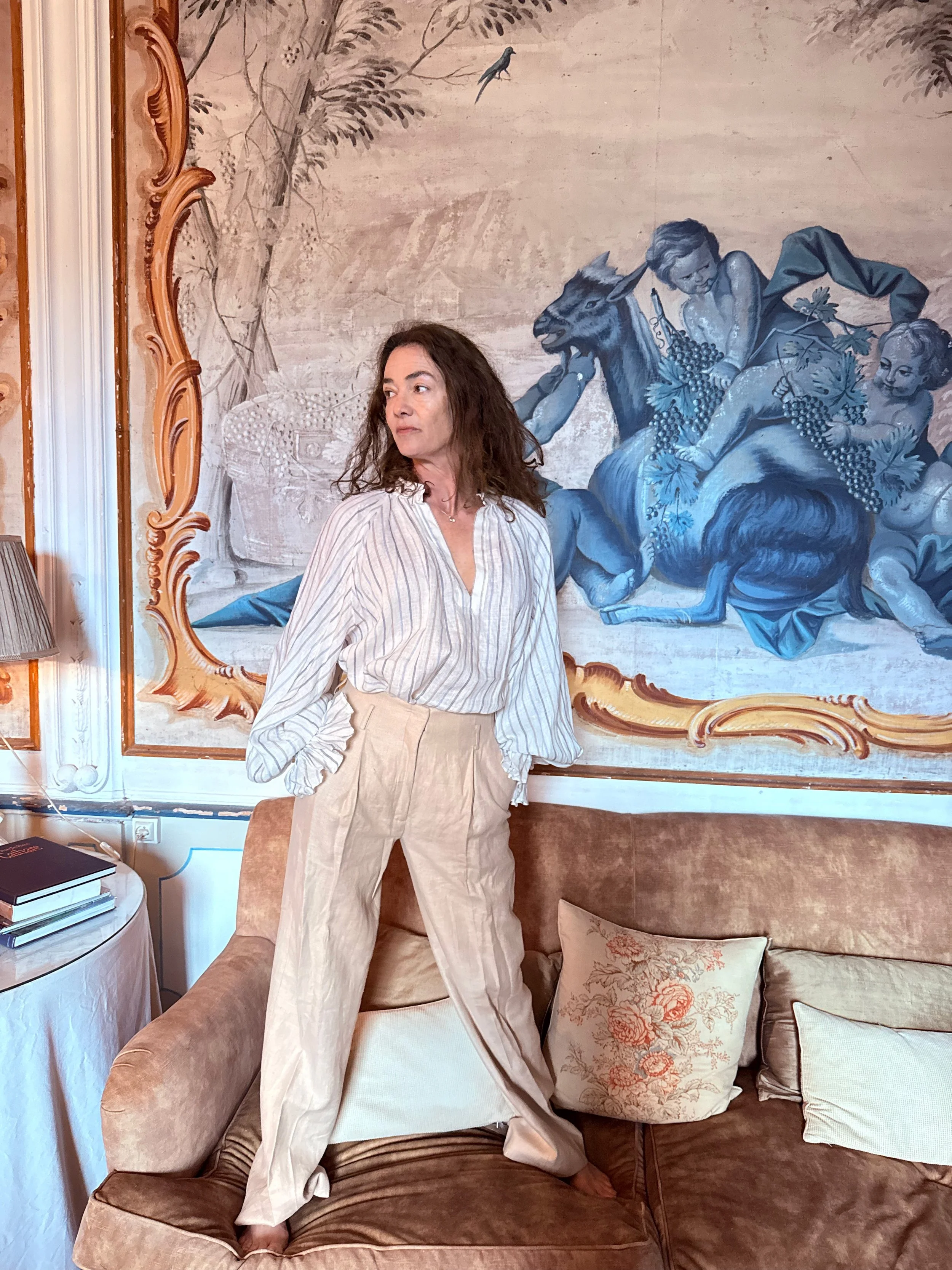Woman with wavy brown hair sitting on a tan sofa, wearing a white and beige striped shirt and beige pants, in a room with a wall mural of a mythological scene featuring a woman, a goat with a human face and a child's face, a bird, and foliage, with p