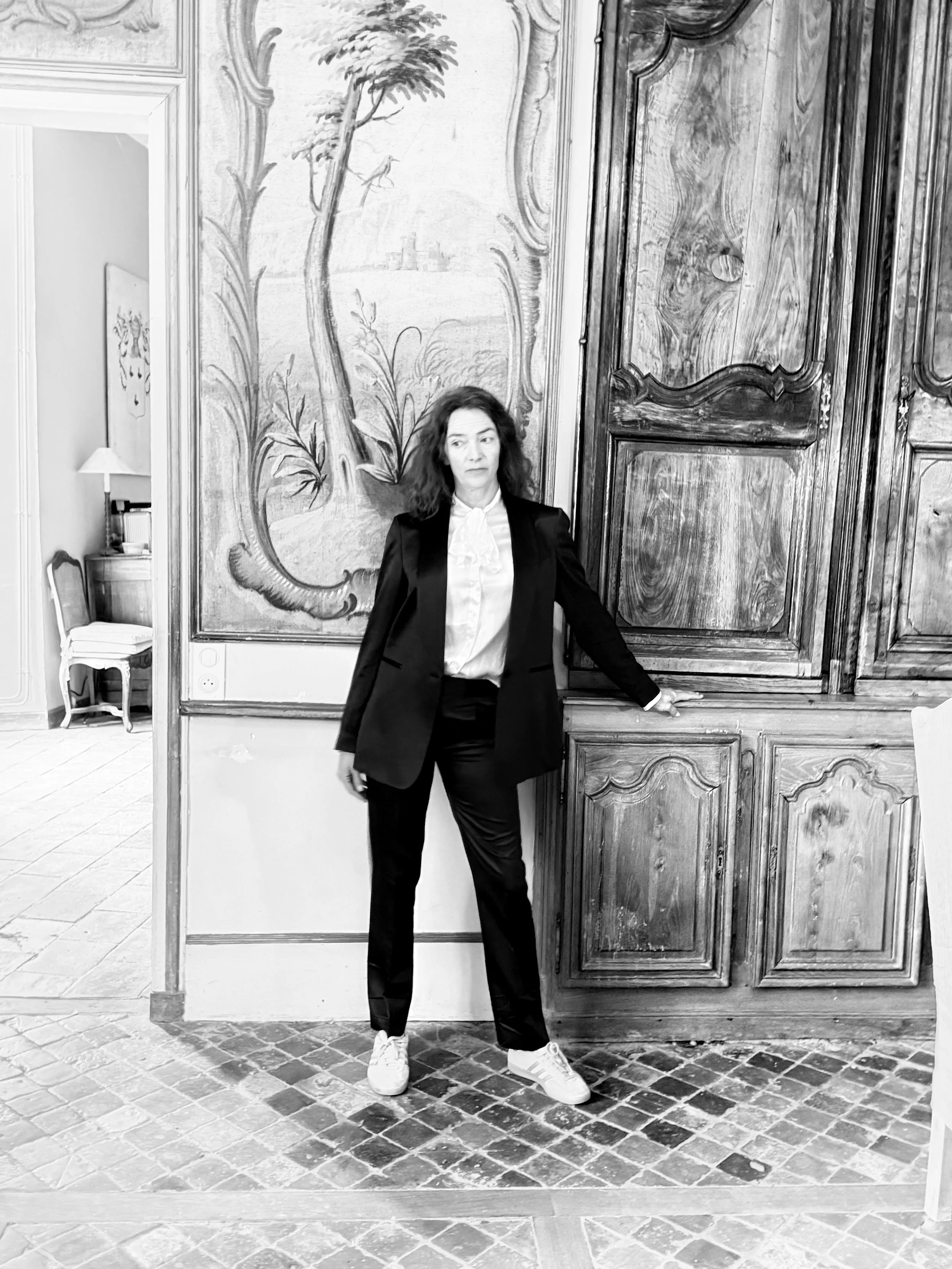 A woman with dark curly hair wearing a black blazer, white shirt, black pants, and white sneakers standing indoors in front of a large wooden cabinet with a painted mural of a tree and bird on the wall behind her.