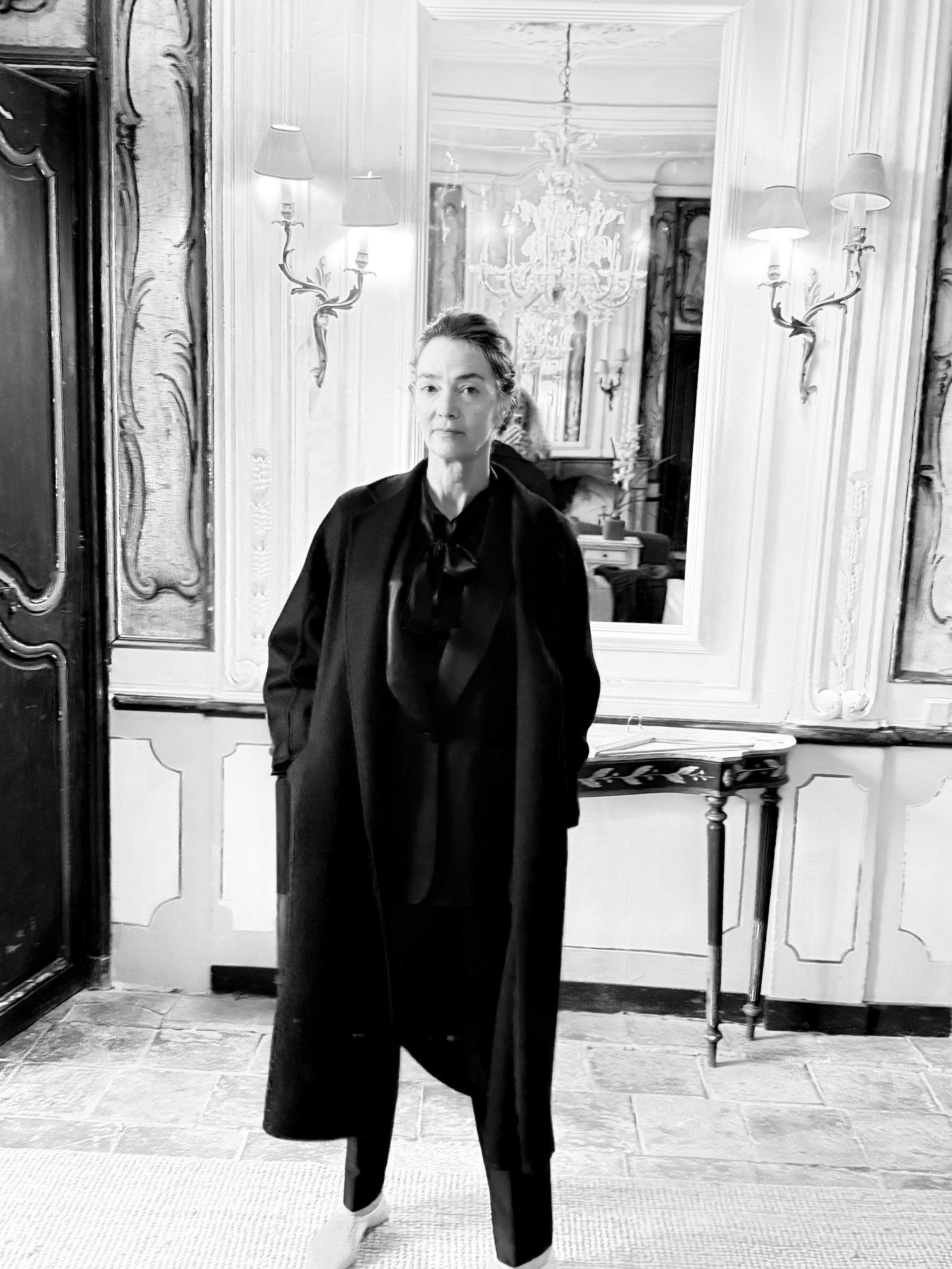 A woman with short dark hair wearing a dark blazer and pants, standing in front of a large mirror in an elegant room with ornate woodwork and a chandelier.