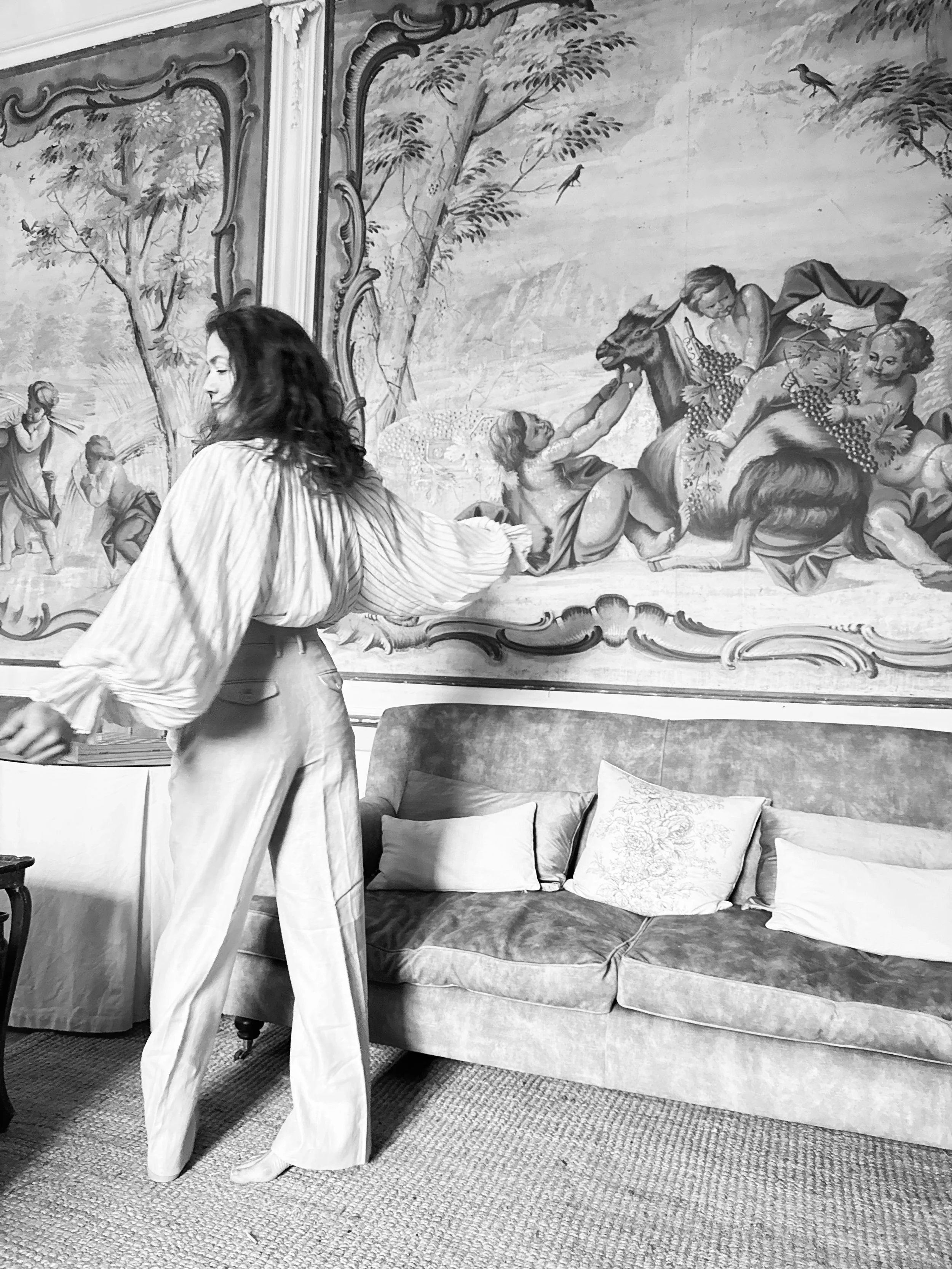 A woman with dark hair wearing a striped blouse and light-colored wide-leg pants walking past a vintage-style sofa with embroidered pillows, in front of a large ornate framed tapestry depicting a pastoral scene with a woman, children, animals, trees,