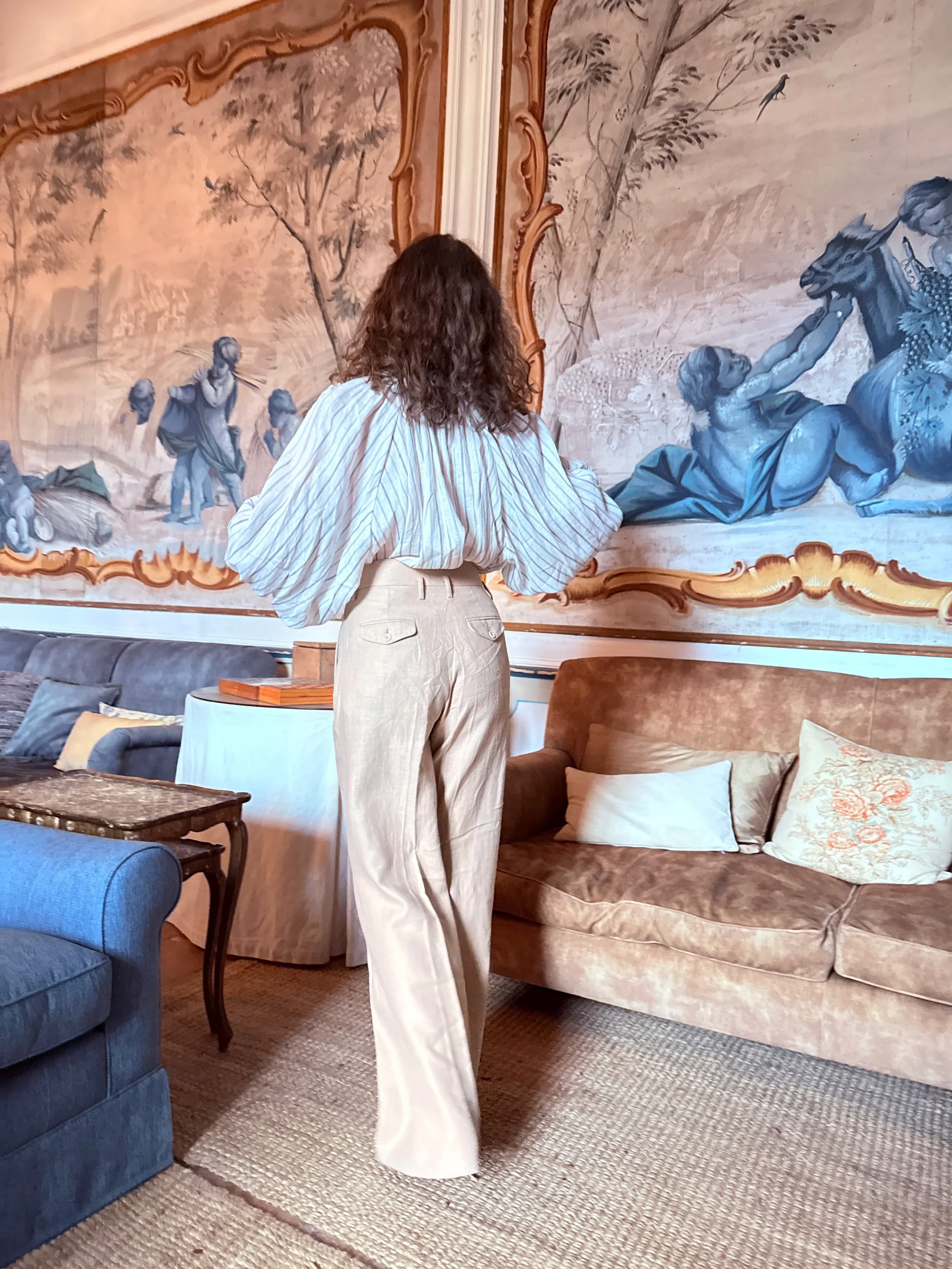 Person with curly hair wearing a striped blouse and beige pants standing in a living room with various sofas, a rug, and a large wall painting depicting a scene with people and nature.