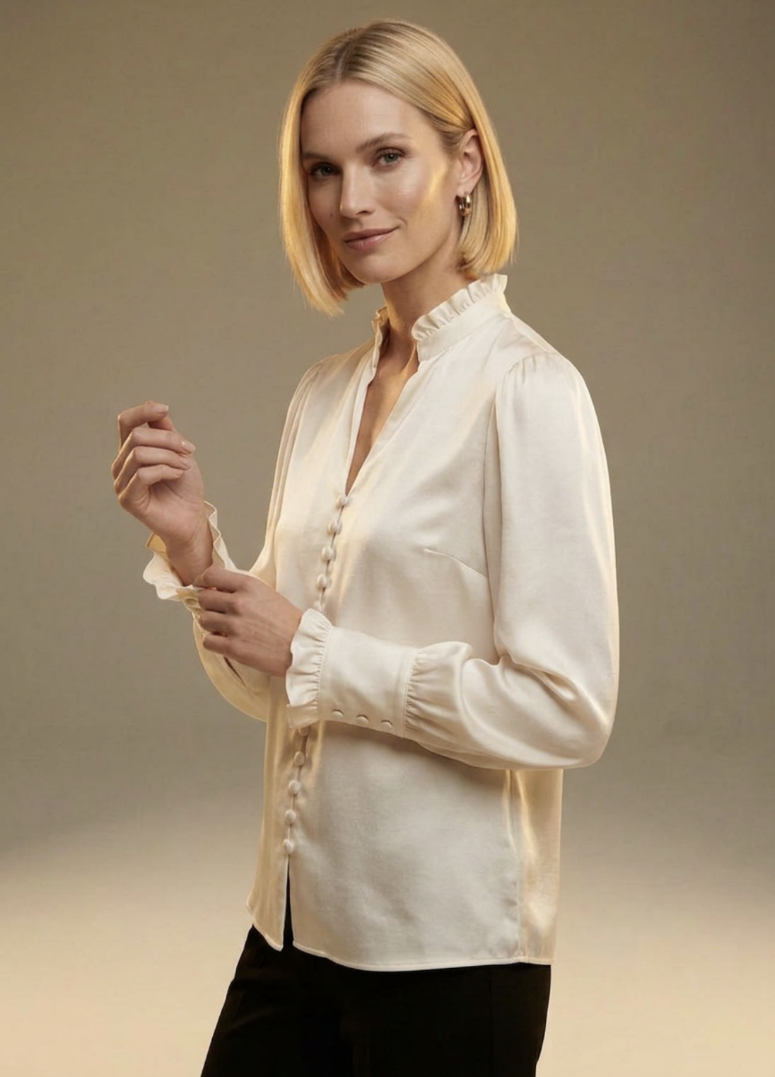 A woman with short blonde hair wearing a cream-colored blouse and black pants, standing against a plain background.