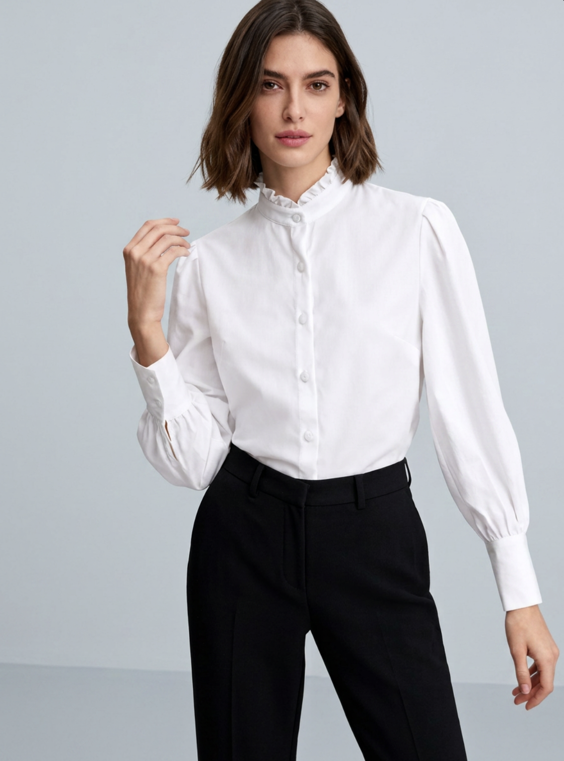 A woman with shoulder-length brown hair wearing a white button-up shirt with a ruffled collar and black trousers.