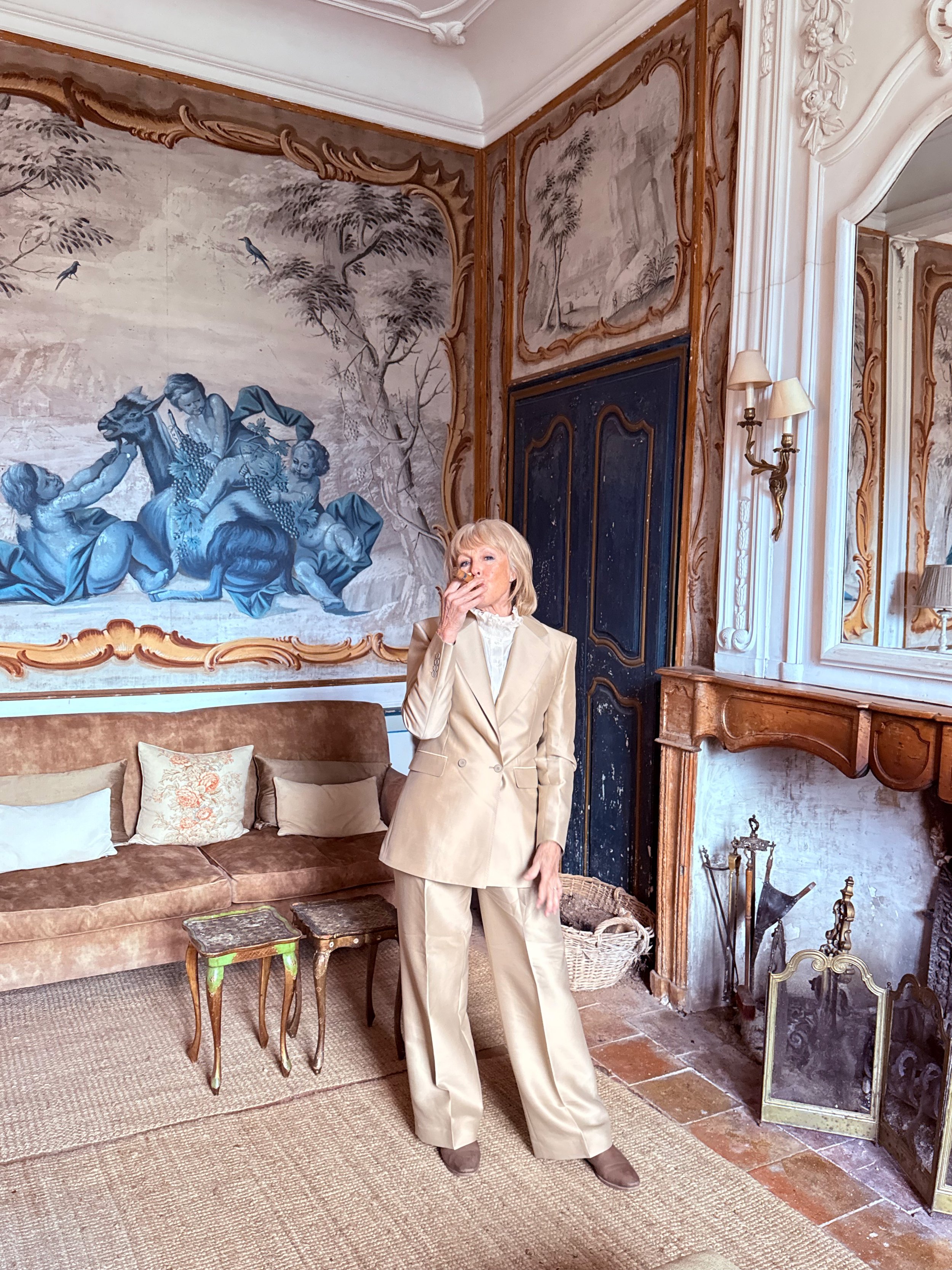 A woman in a beige suit standing in a room with ornate decor, including a large wall mural, wooden furniture, and a fireplace.