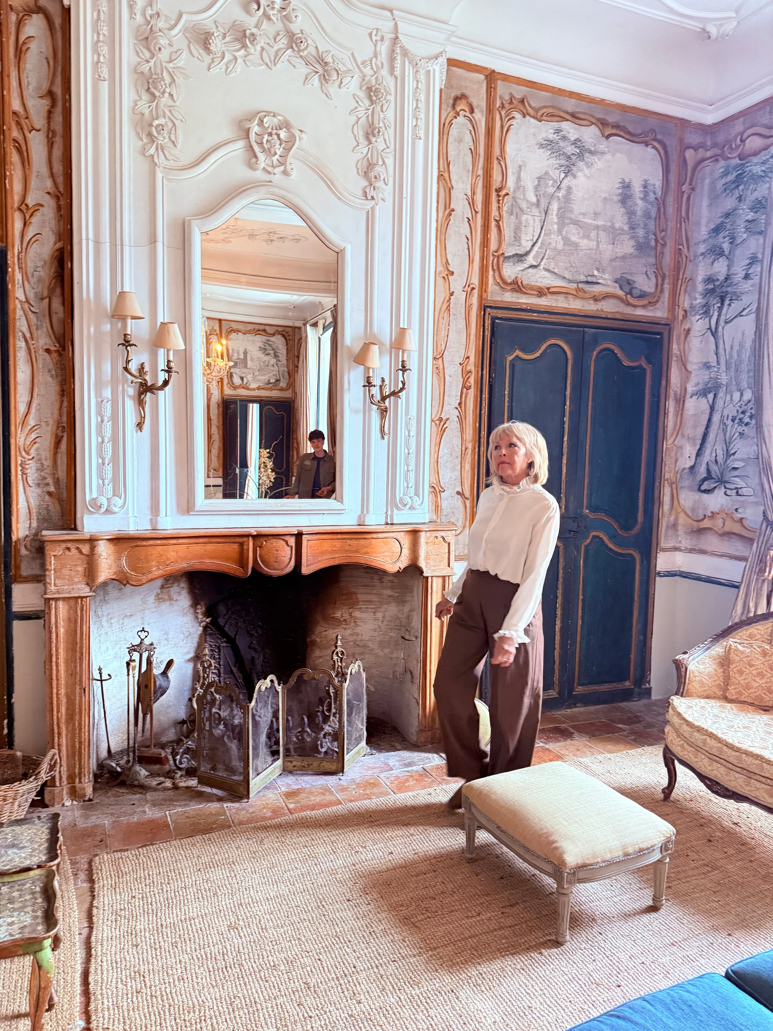 A woman with blond hair and a white blouse walks in an elegant living room with ornate wood and plasterwork. The room features a large mirror above a fireplace, a tapestry on the wall, and vintage furniture including a bench, an armchair, and a firep