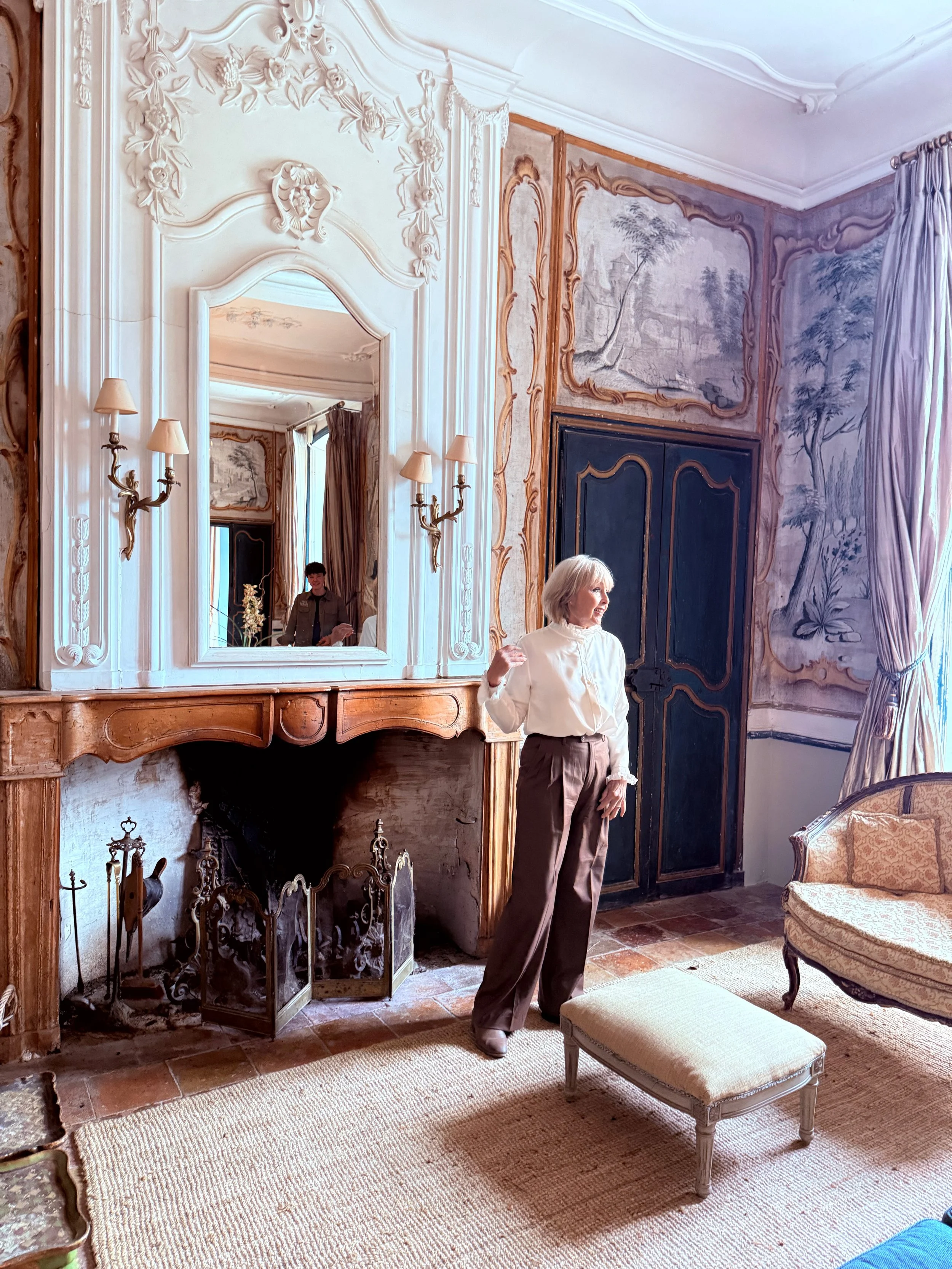 A woman with gray hair wearing a white blouse and brown pants standing in a room with ornate wall decorations, a large mirror, a fireplace, and antique furniture. There is a reflection of a person in the mirror.
