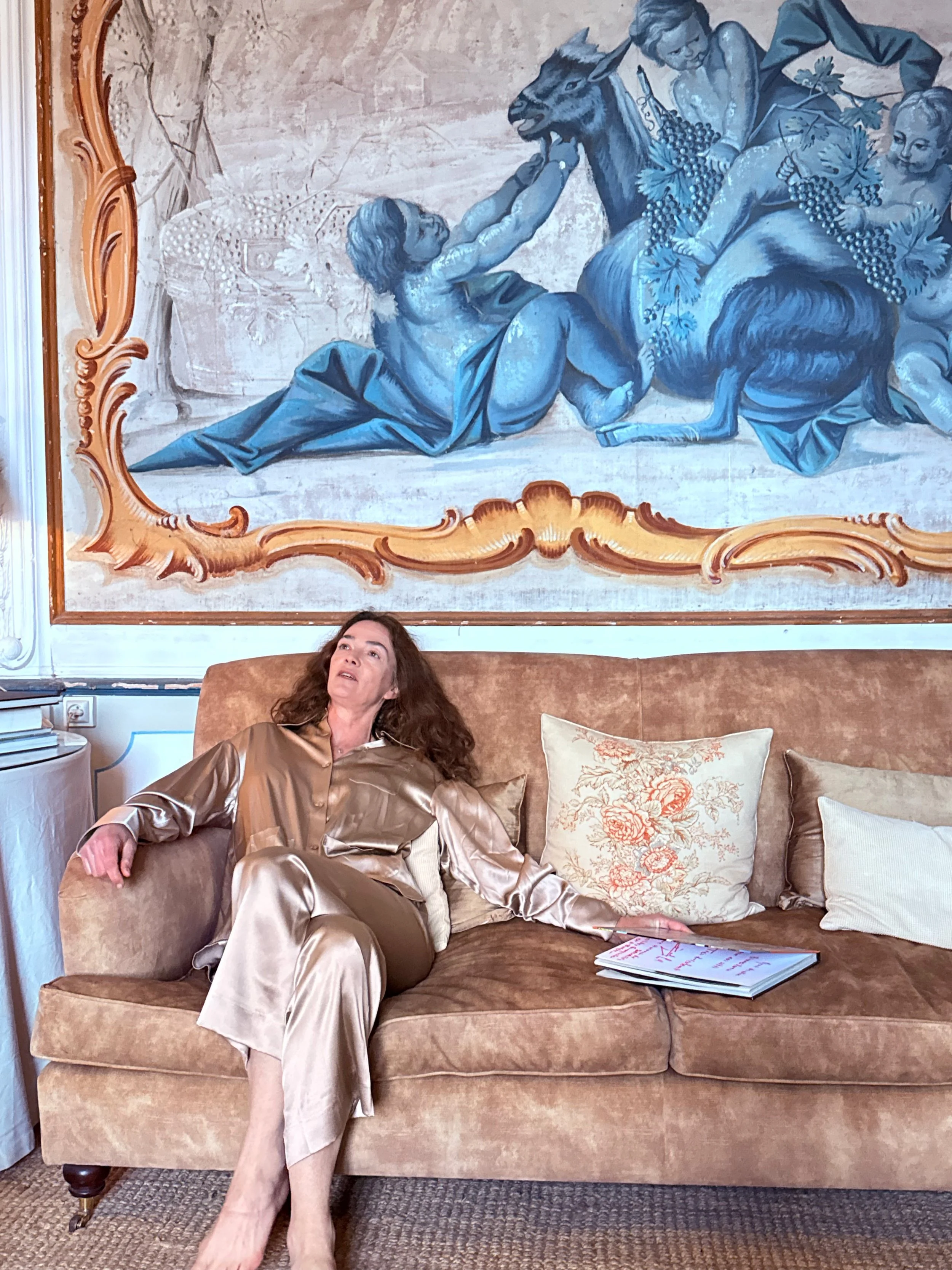 A woman in shiny gold pajamas sitting on a beige sofa, leaning back with her legs crossed at the ankle. There are decorative pillows on the sofa, and a large ornate framed wall art depicting children playing with a goat hangs behind her.