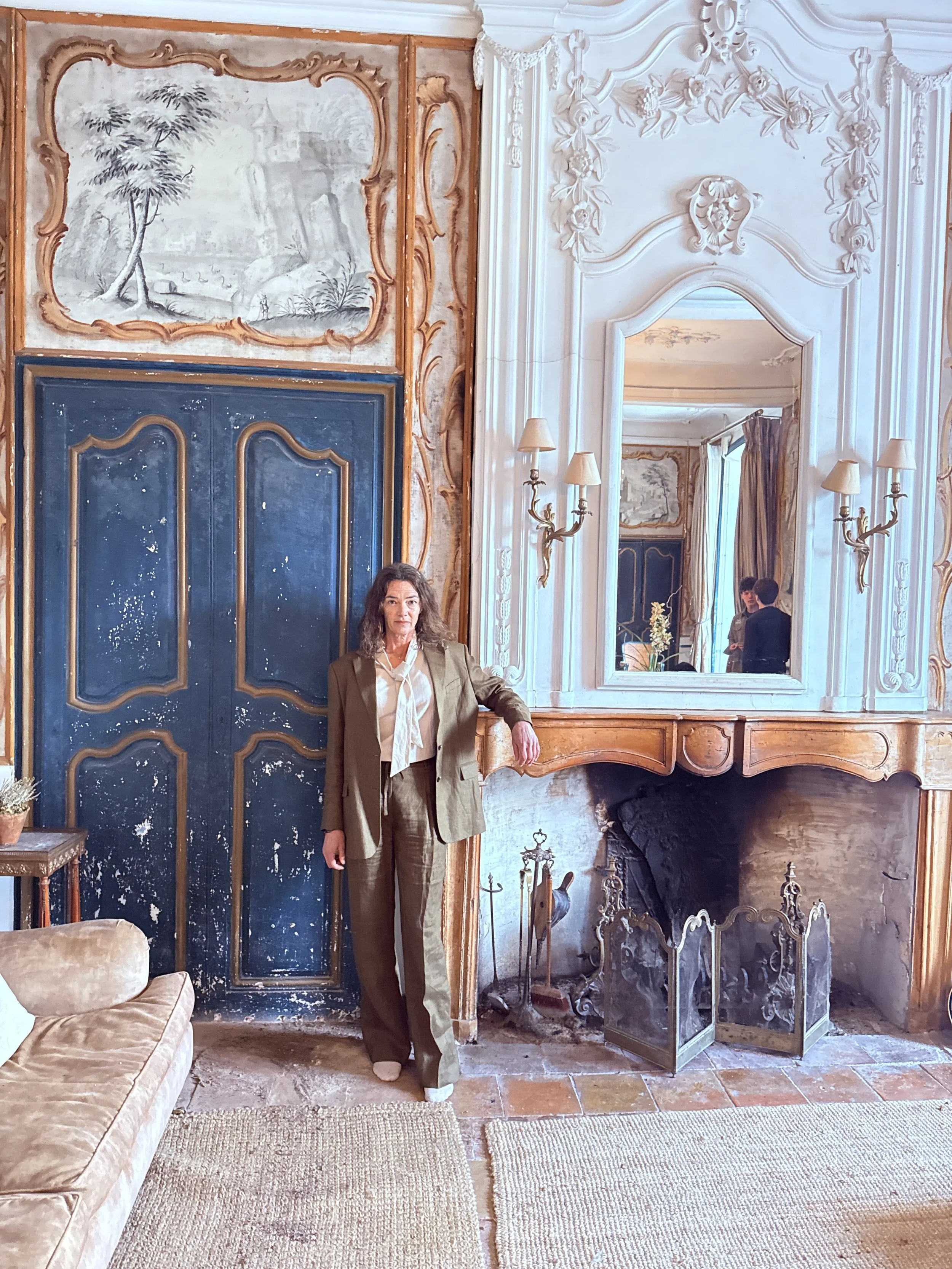 Woman in brown suit standing in a richly decorated room with ornate white walls, large mirror, and a fireplace, capturing her reflection in the mirror.