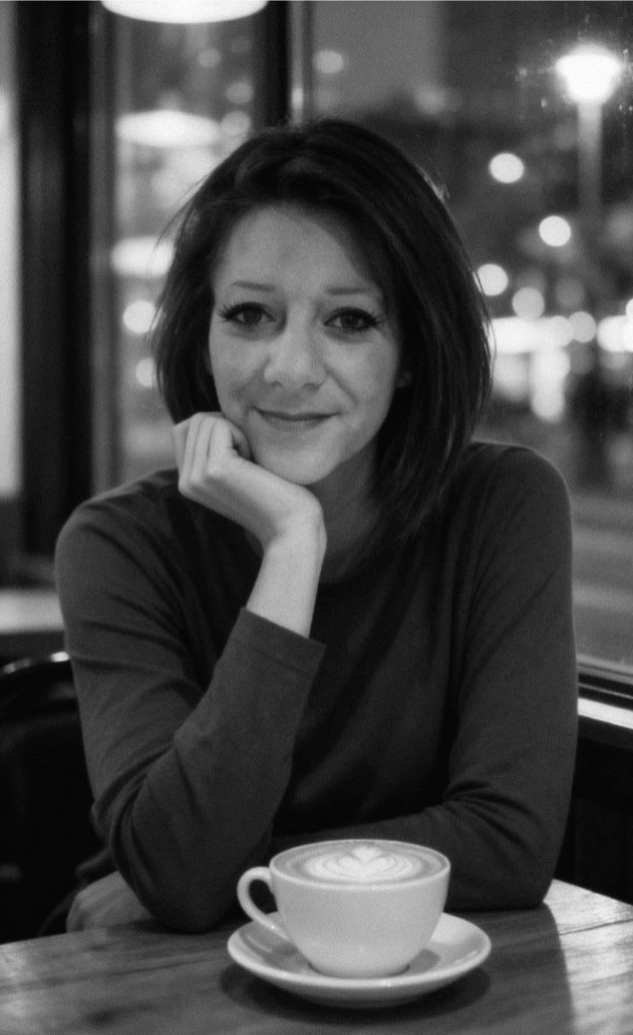 Une femme assise à une table dans un café, tenant son visage avec une main, avec une tasse de café devant elle, dans un décor de café avec des lumières floues à l'arrière-plan.