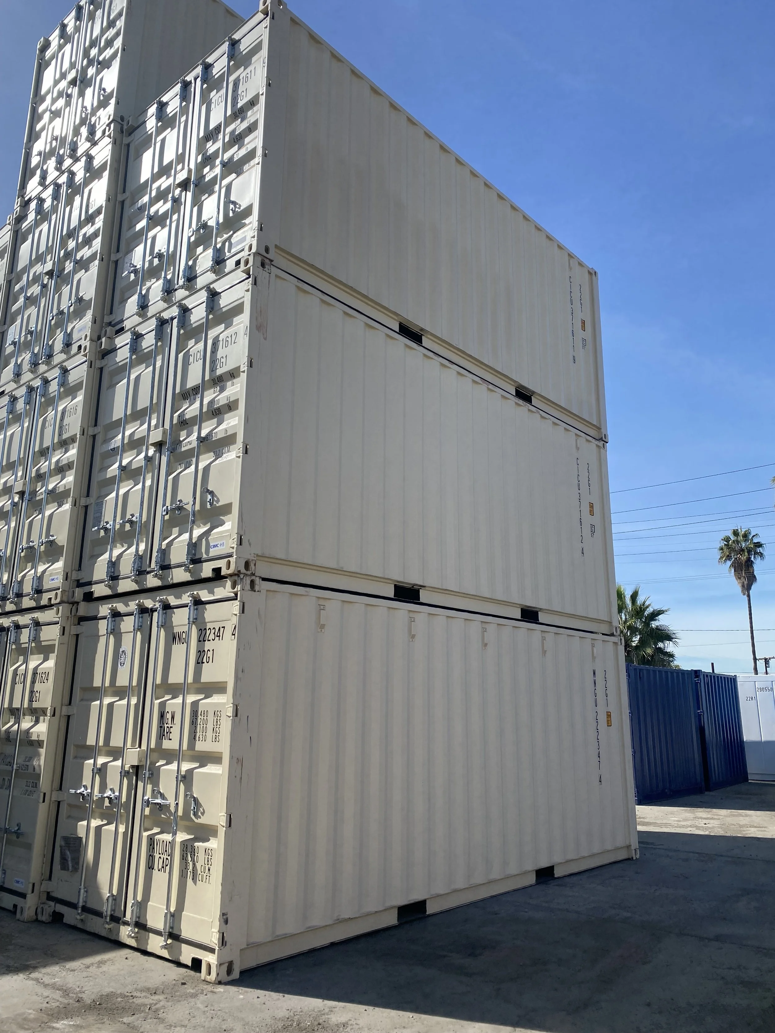 Stacked white shipping containers outdoors under a clear blue sky, with palm trees and utility lines visible in the background.