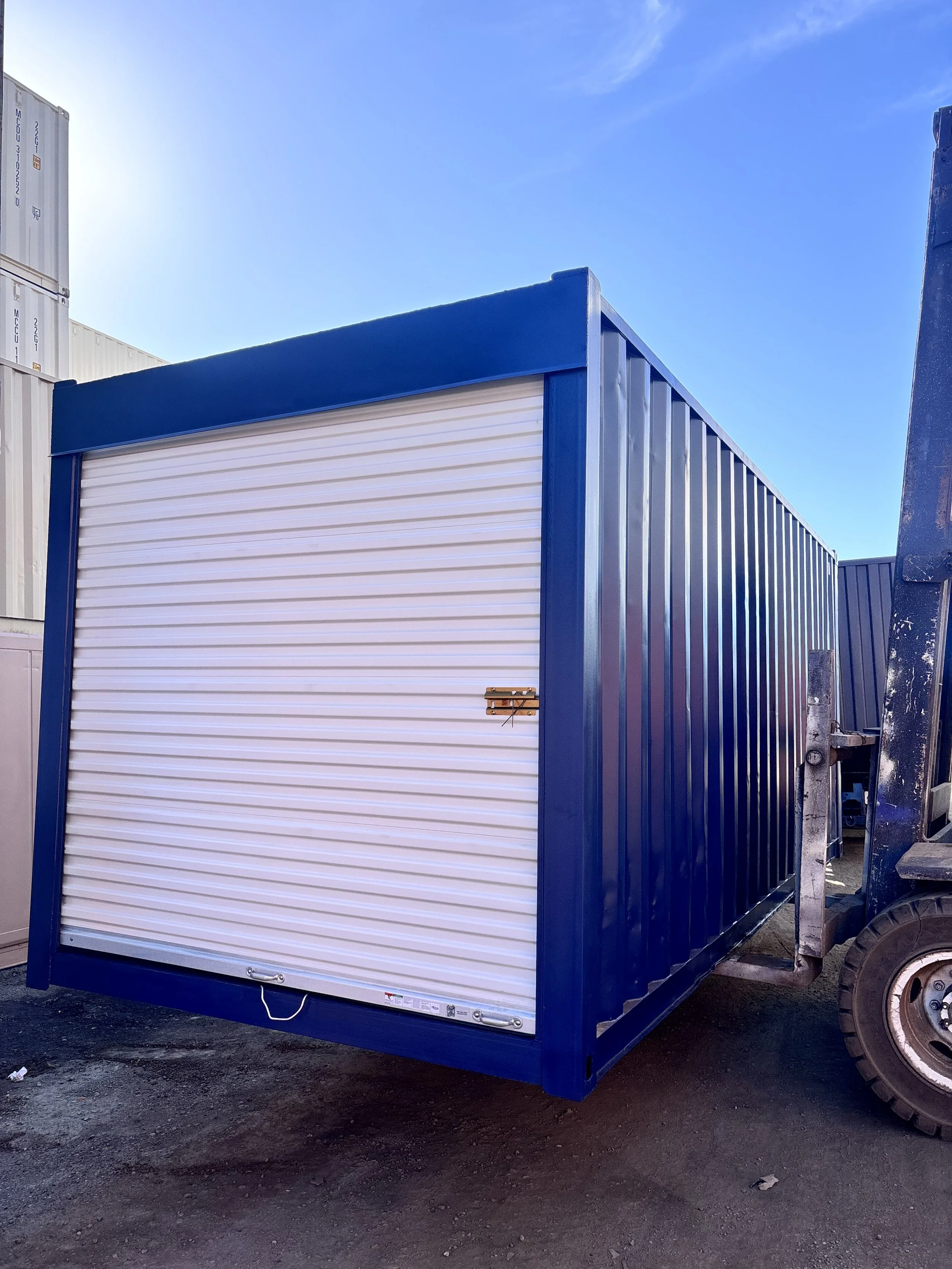 A blue 20ft refurbished wind and wateright storage container with a white roll-up door, situated outdoors on a dirt surface, with clear blue sky overhead.