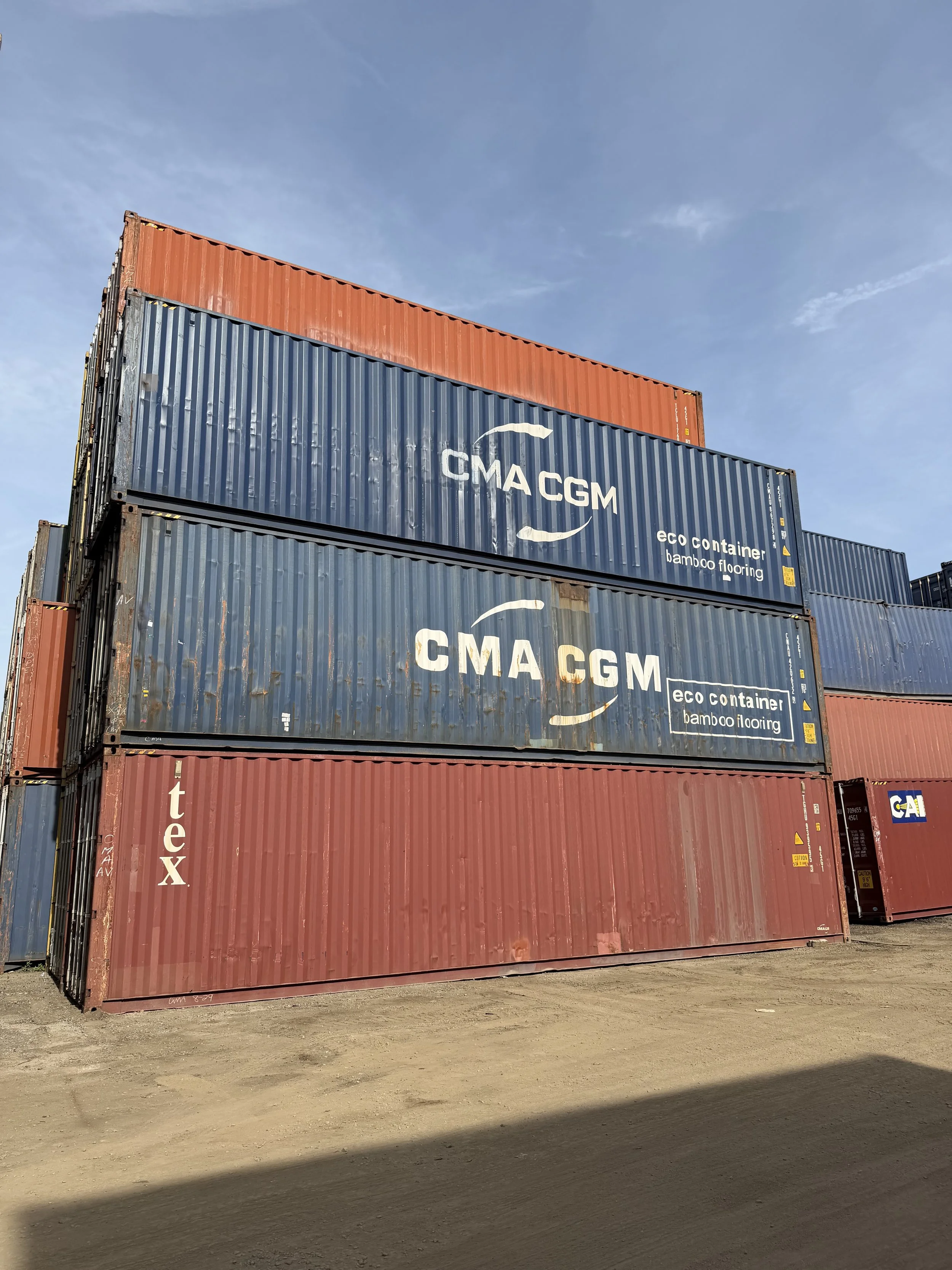 Stack of 40ft used wind and watertight shipping containers, blue and red, with logos and text, under a clear blue sky.