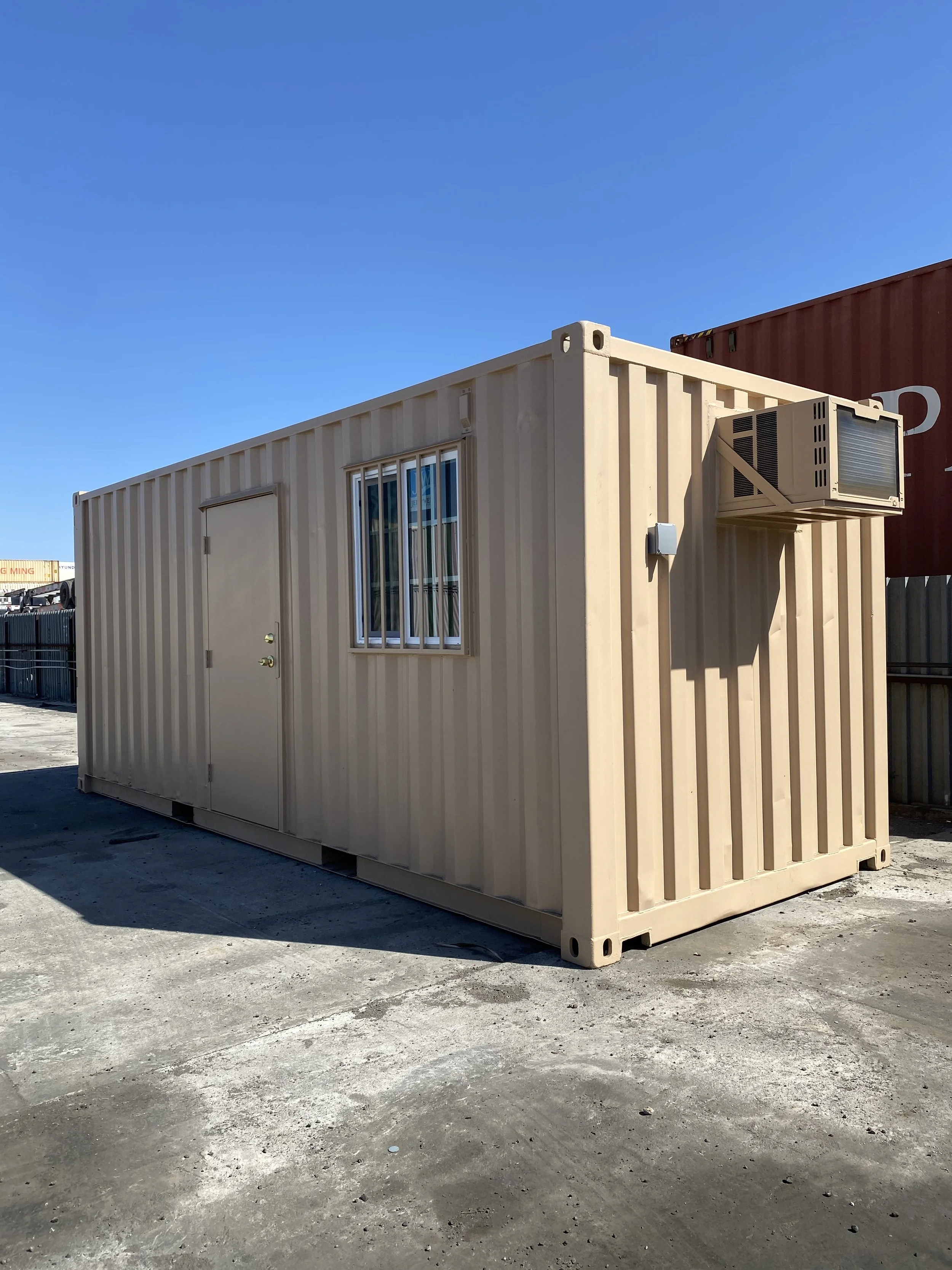 A beige portable building with a door, a barred window, and an air conditioning unit on the side, situated outdoors under a clear blue sky.