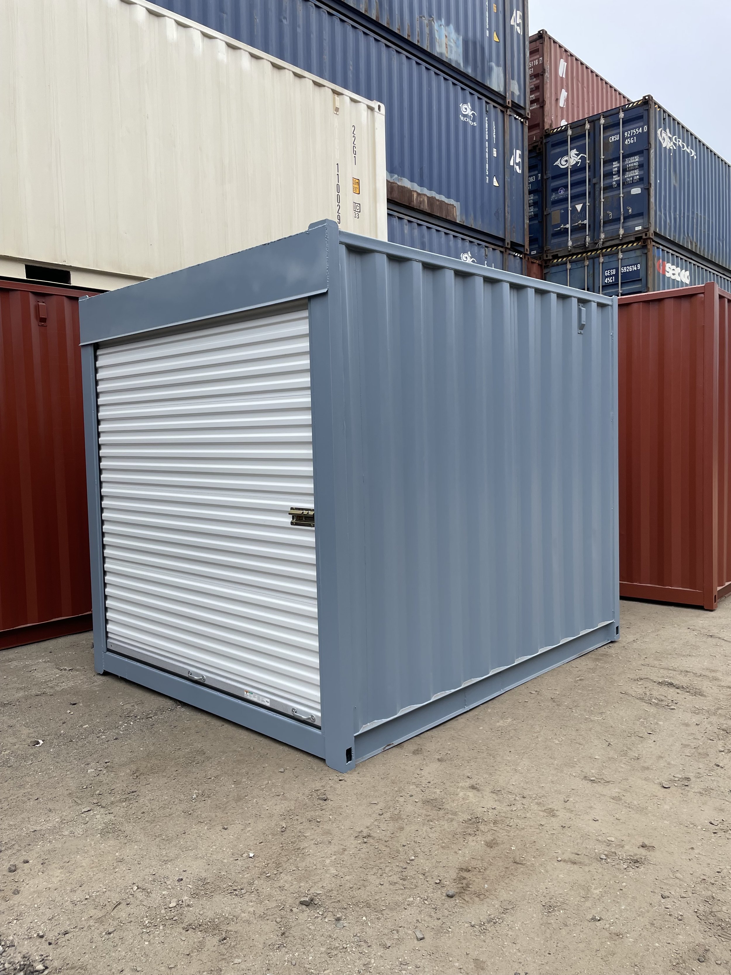 Blue shipping container with a white roll-up door, situated on dirt ground, surrounded by other red and blue shipping containers stacked in the background.