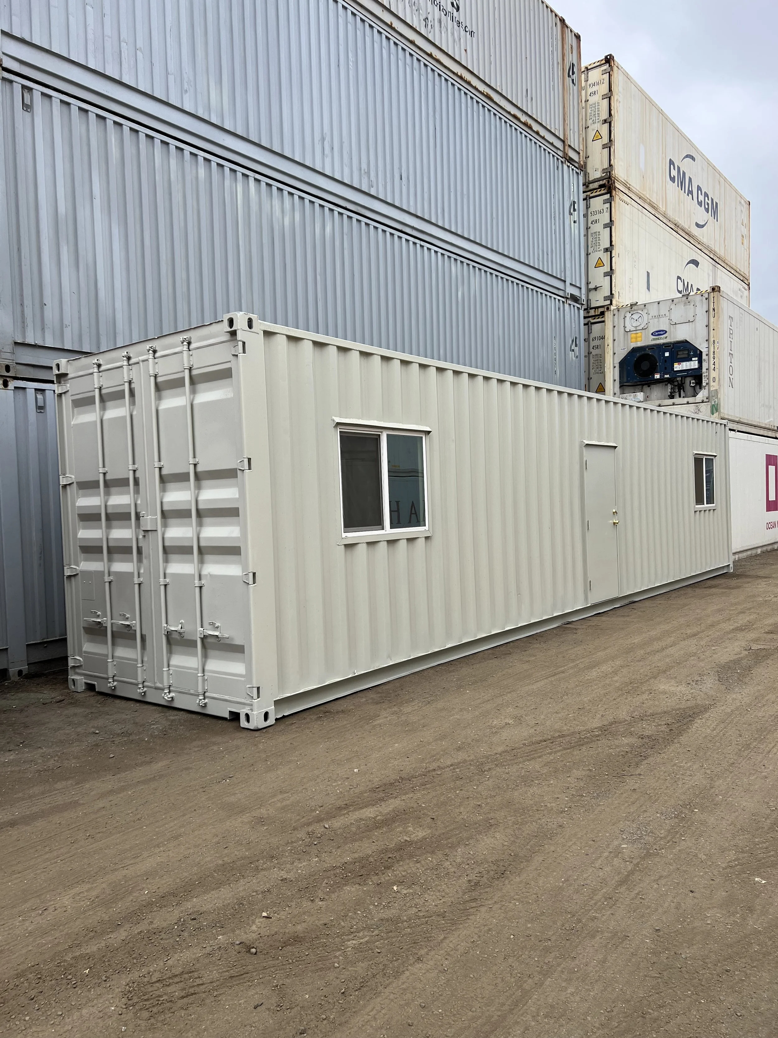 A white shipping container with windows and a door placed on dirt ground, stacked with large shipping containers in a storage yard.