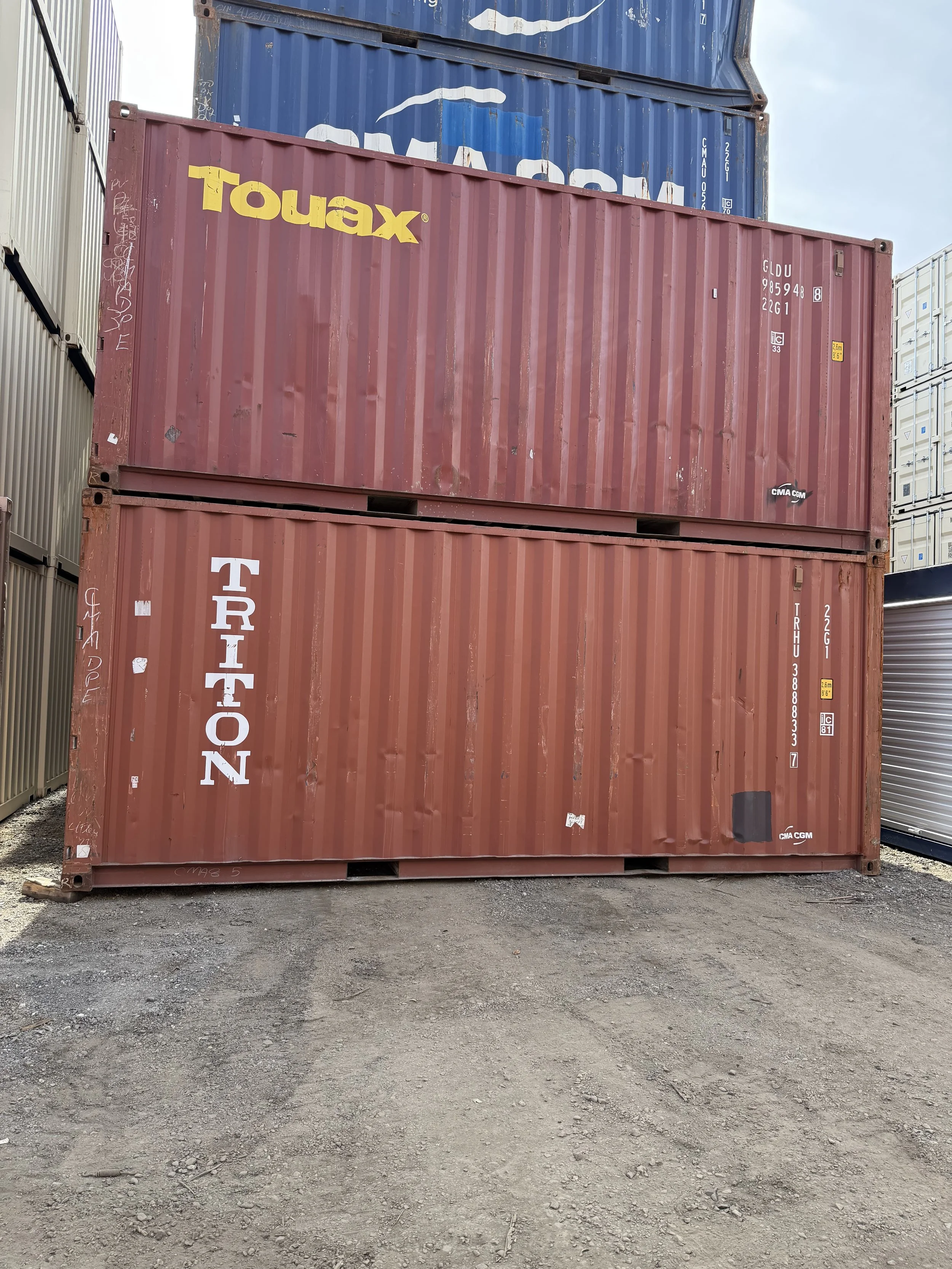 Two stacked used 20ft wind and watertight shipping containers in an outdoor storage yard, with one container labeled 'TRITON' and the other 'touax', surrounded by other containers.