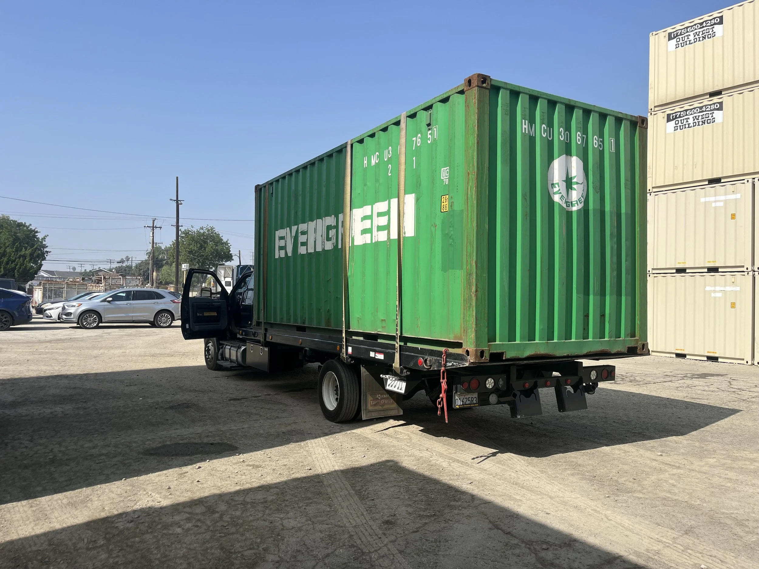 20ft used wind and watertight cargo worth Green shipping container on a flatbed truck parked on a dirt lot, with cars and stacked beige shipping containers in the background on a clear day.