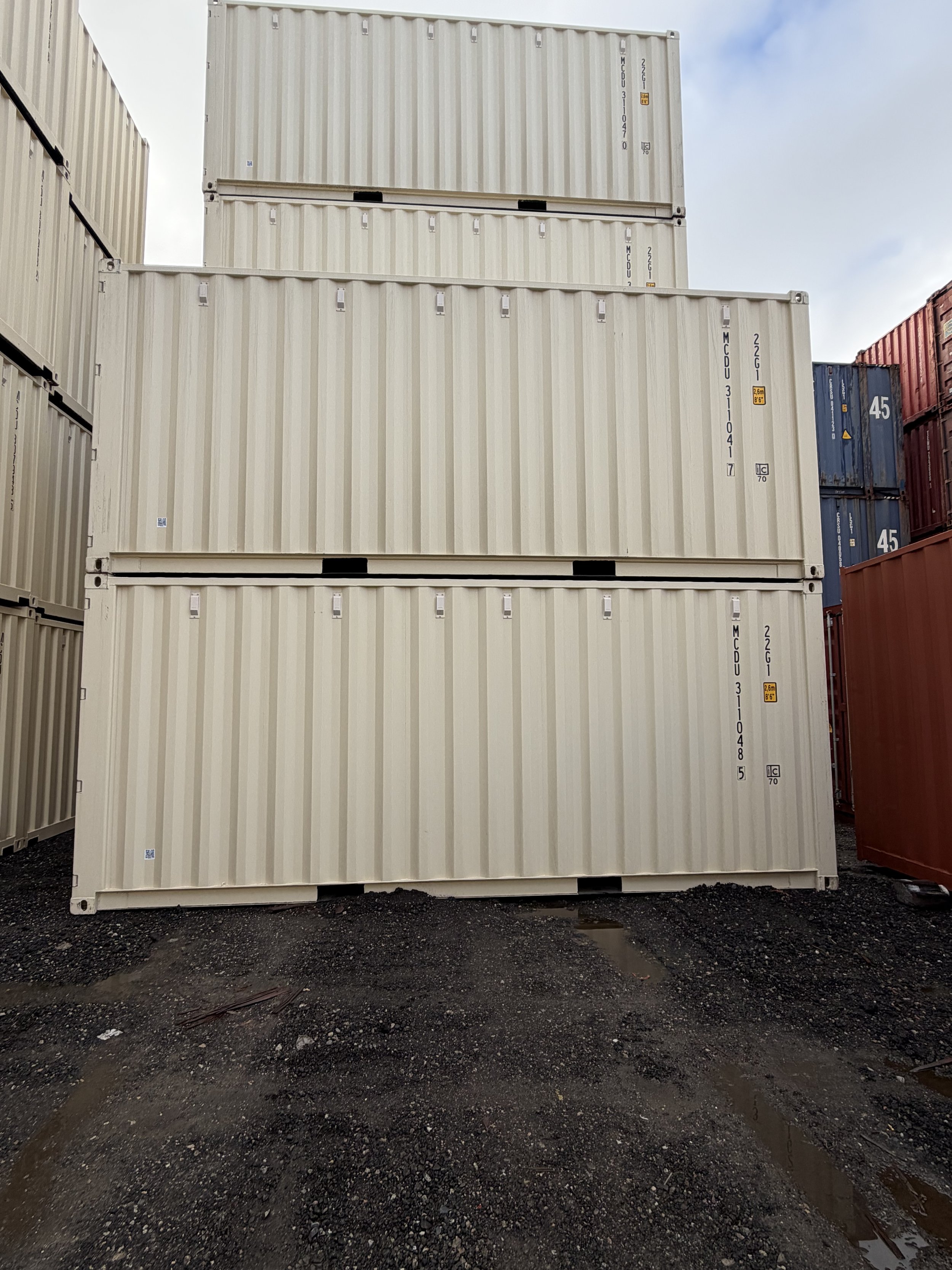 Three stacked beige 20FT One Trip Wind and Watertight shipping containers in an outdoor storage yard with other containers visible to the sides. 