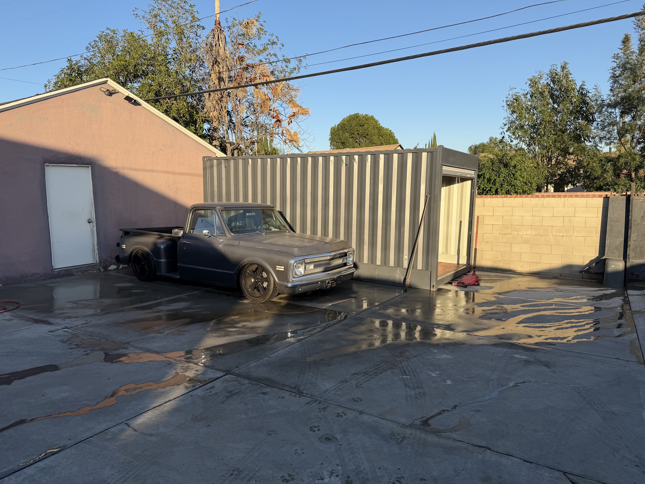 A vintage dark gray pickup truck parked on a wet concrete driveway next to a portable storage container, with a pink building and trees in the background, under a partly cloudy sky.