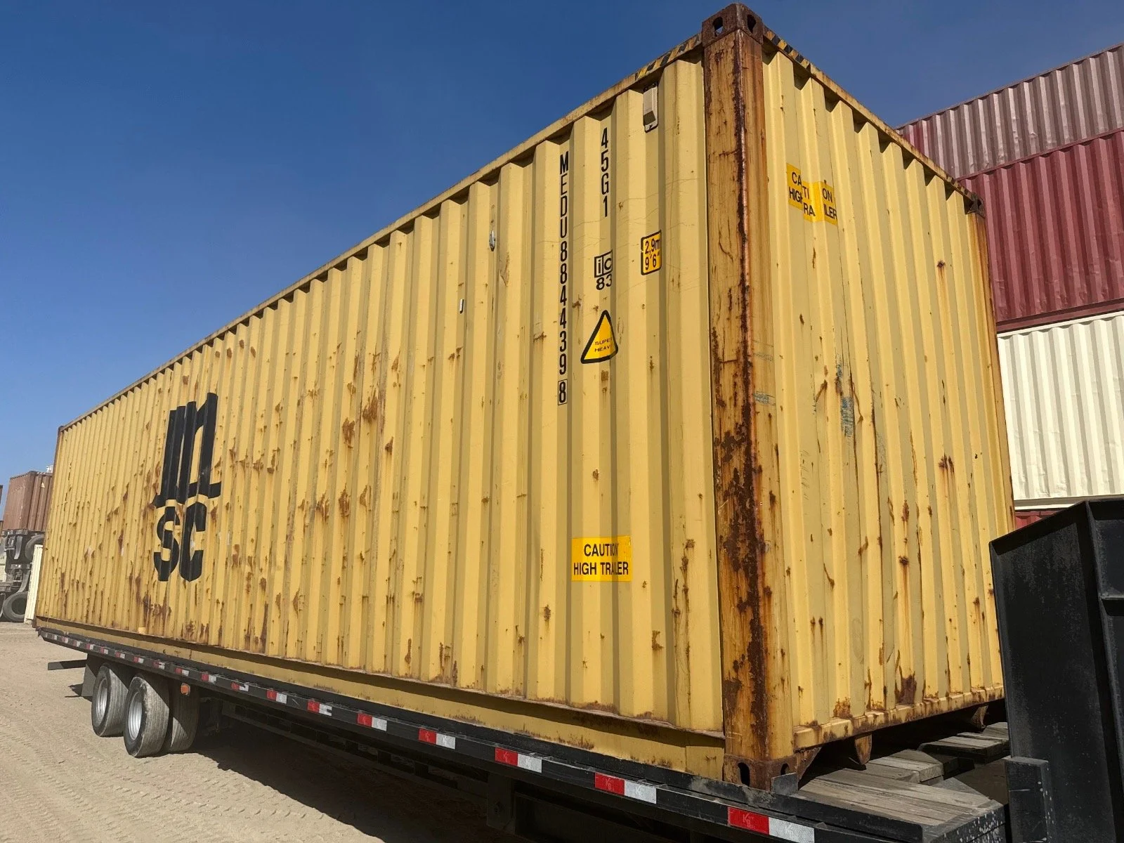 Yellow used 40ft wind and watertight shipping container with rust spots and black lettering, labeled 'ML SC', on a flatbed trailer, with a blue sky background.
