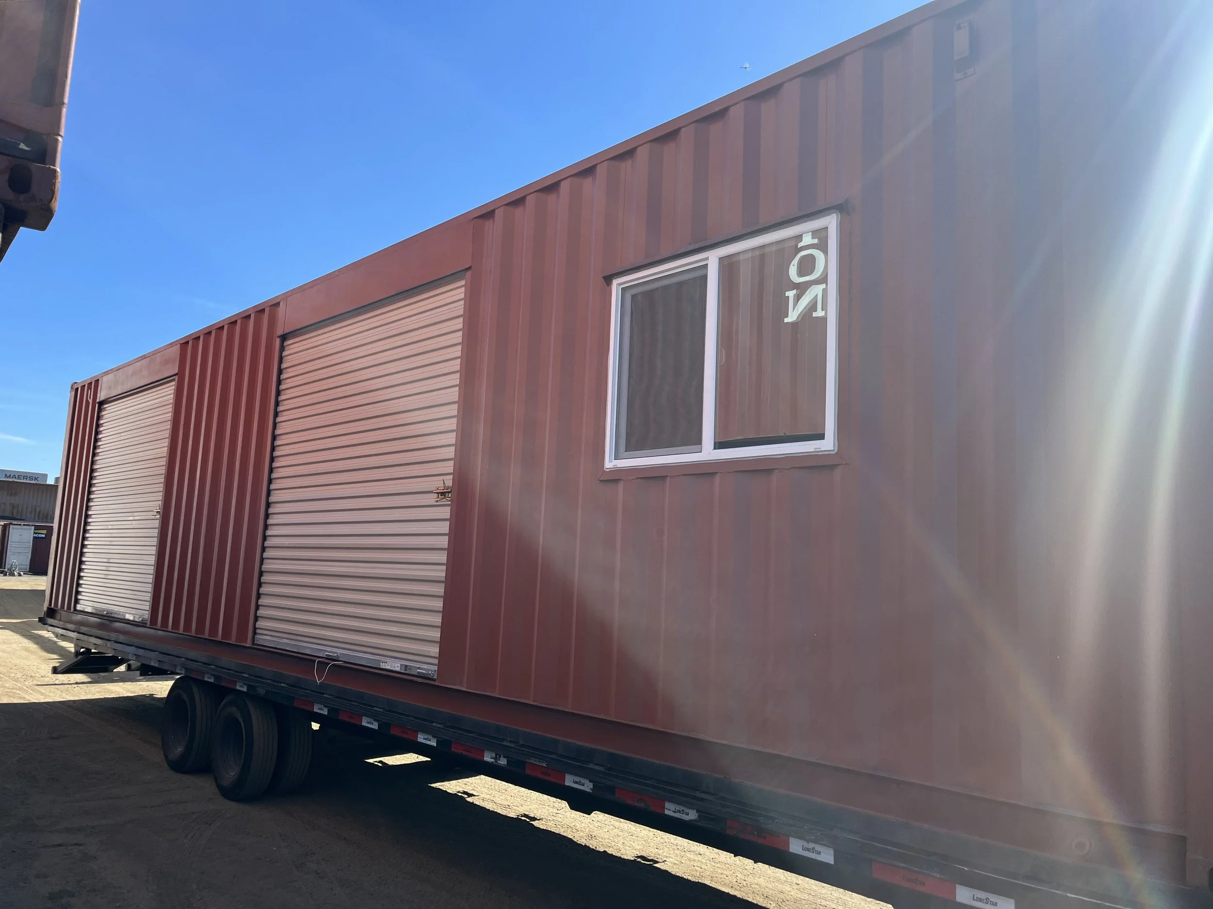 Red modular building with a large rolling door and a window, on a flatbed trailer, under a clear blue sky, with sunlight glare.