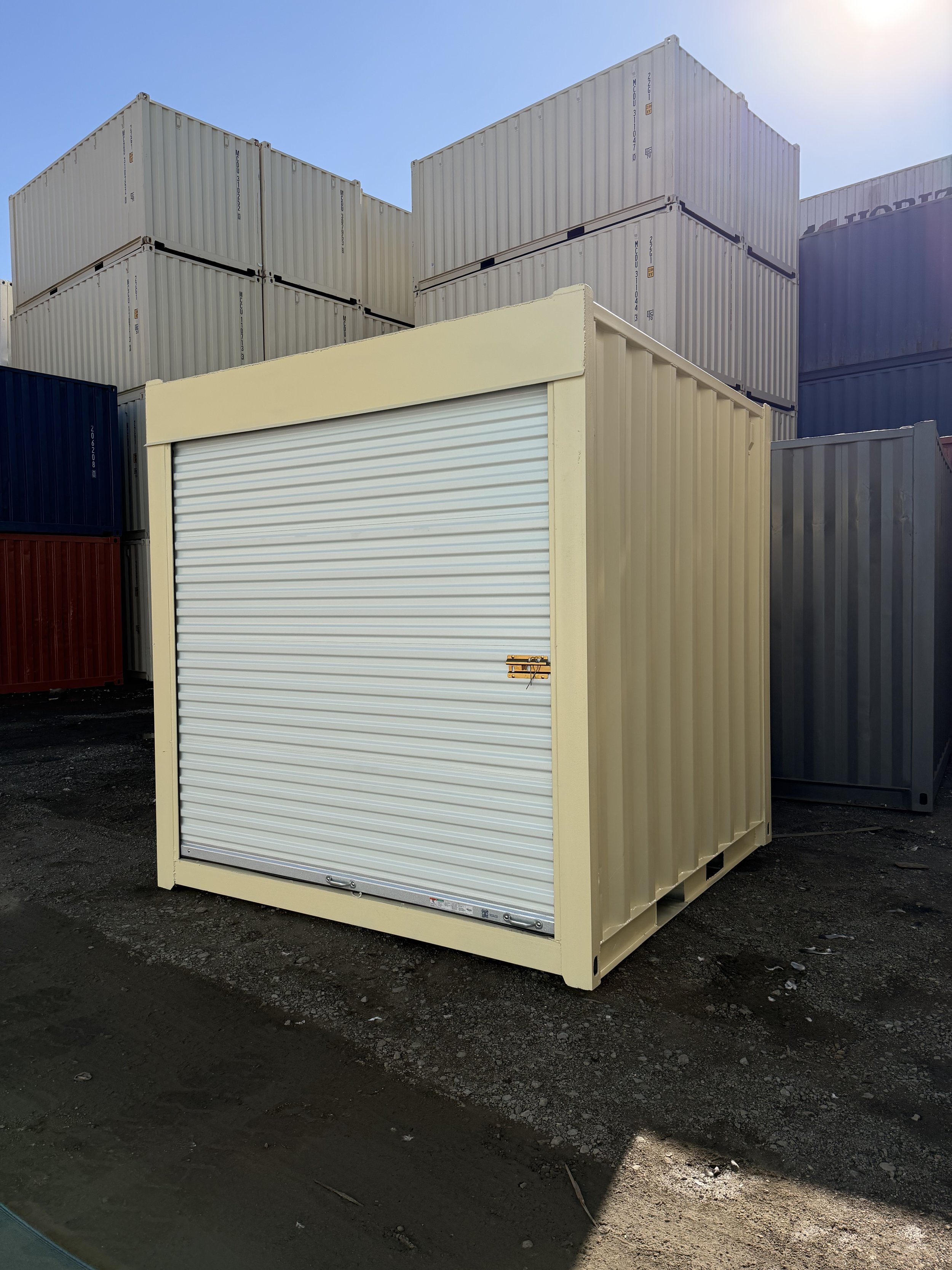 A beige 10FT storage container with a white roll-up door, situated outdoors on a dirt ground, surrounded by stacked shipping containers.