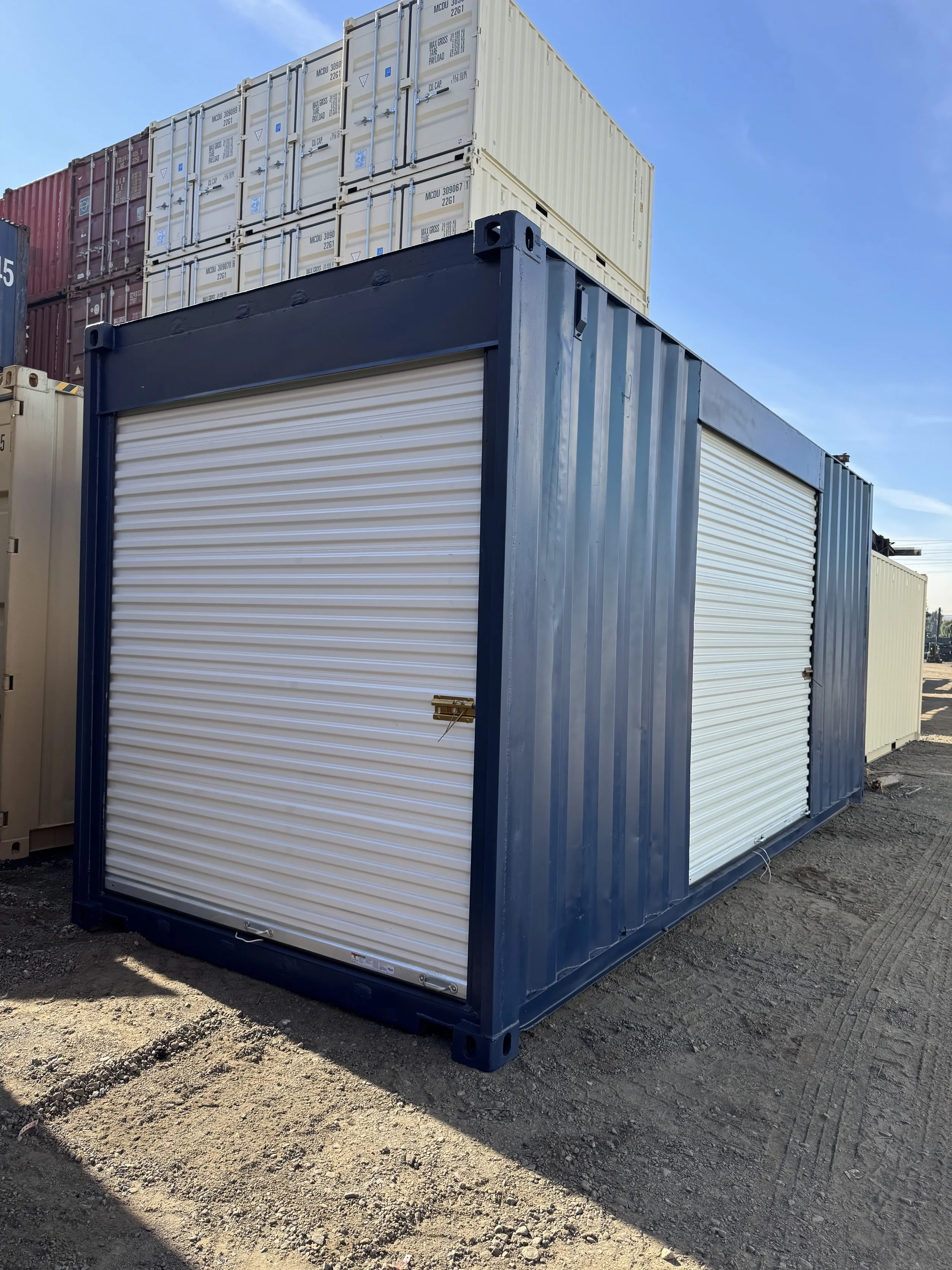 20FT Shipping container with a roll-up door, blue frame, on a dirt surface with stacked containers in the background under a blue sky.