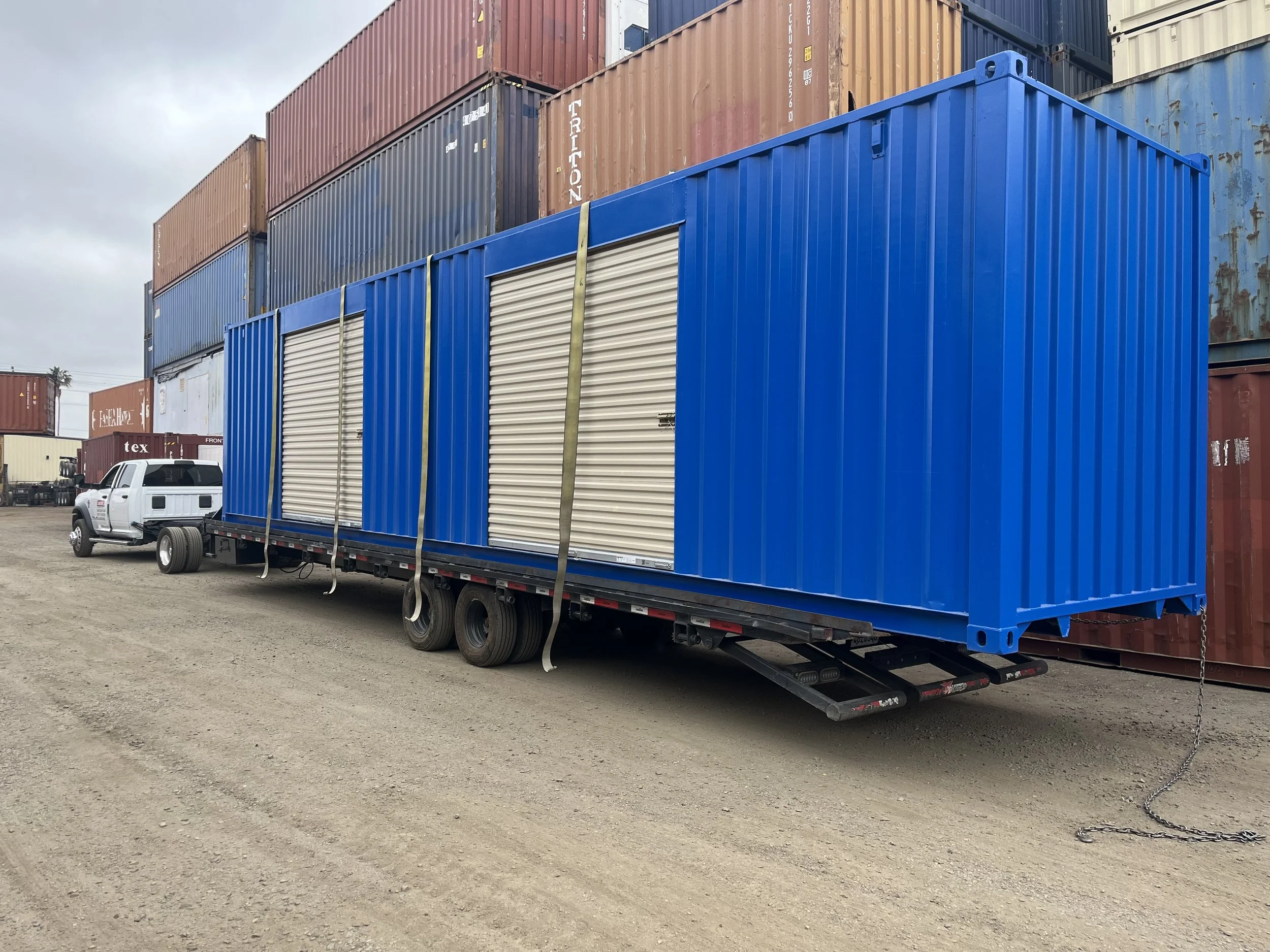A large blue shipping container with roll-up doors on a flatbed trailer, parked among other shipping containers and a white pickup truck in a shipping yard.
