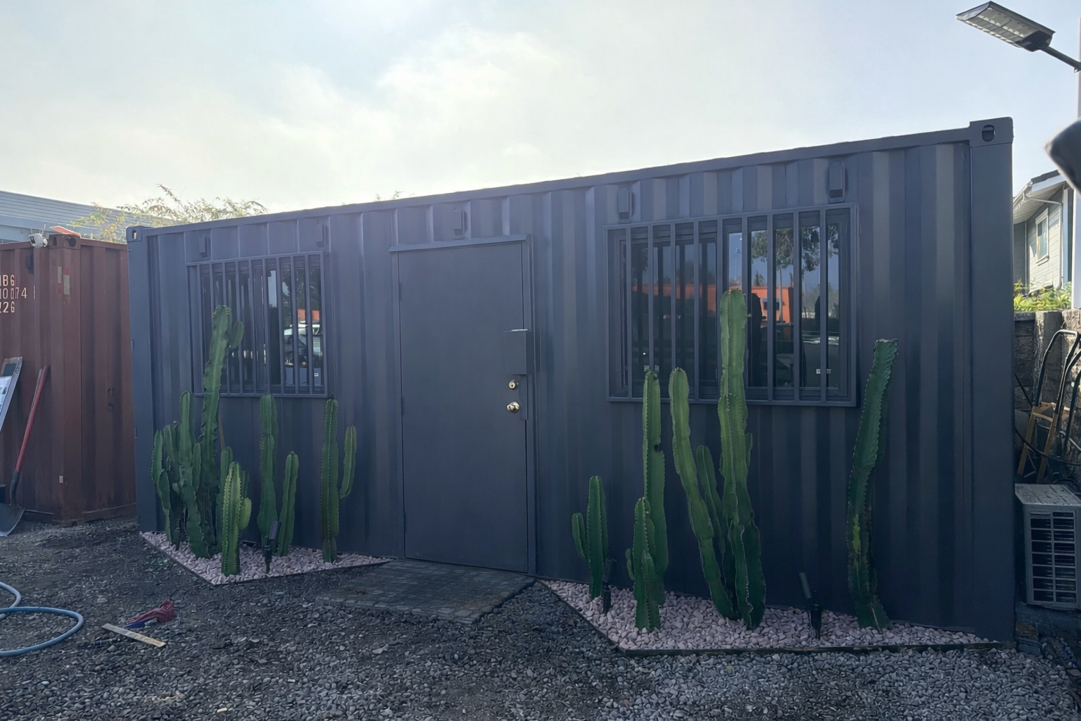 A small dark gray 20ft office made from a shipping container with security bars on the windows and a door, surrounded by tall cacti and gravel ground.