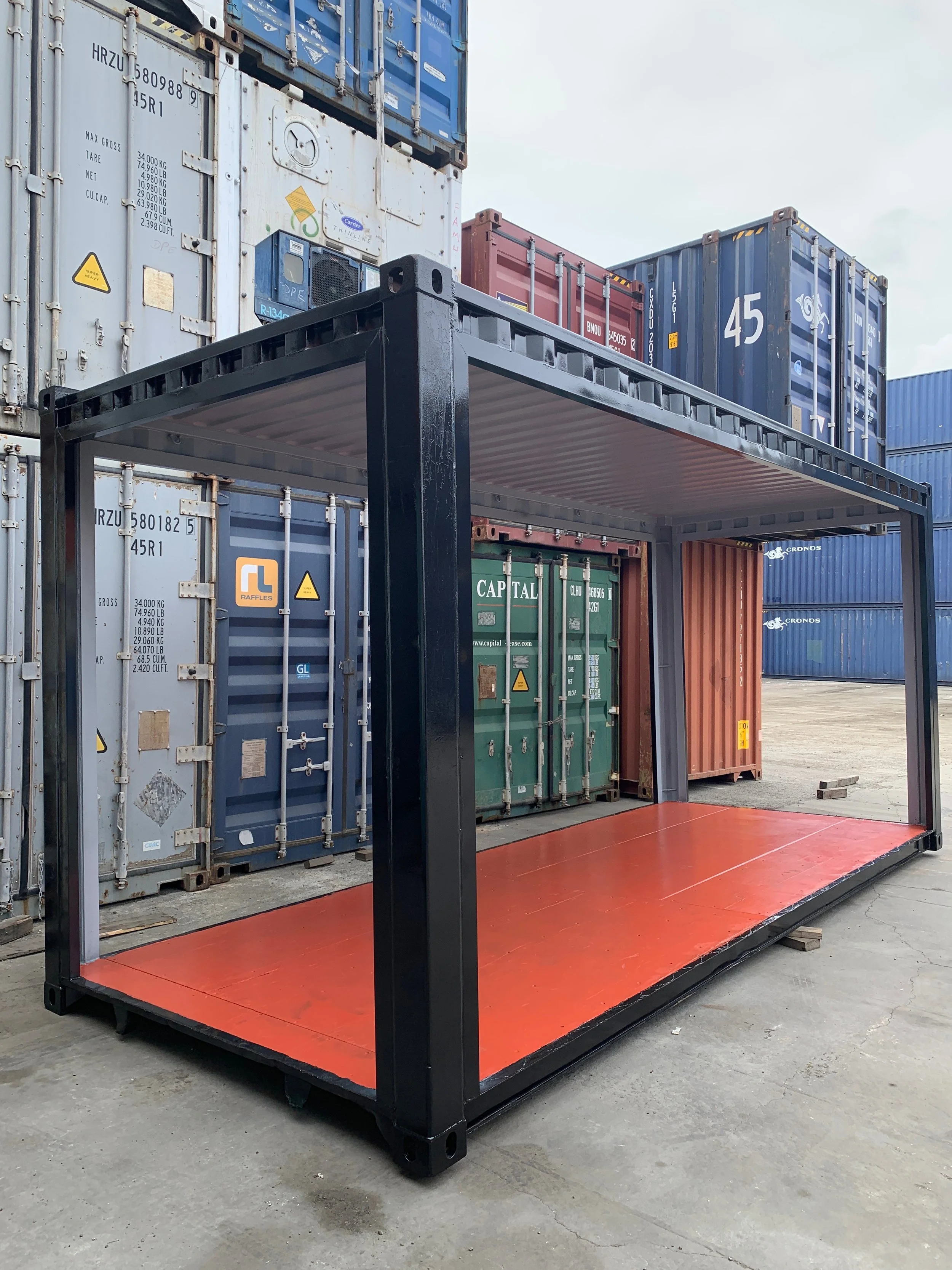 A shipping container frame with a red floor in an outdoor shipping yard, with stacked shipping containers in the background.