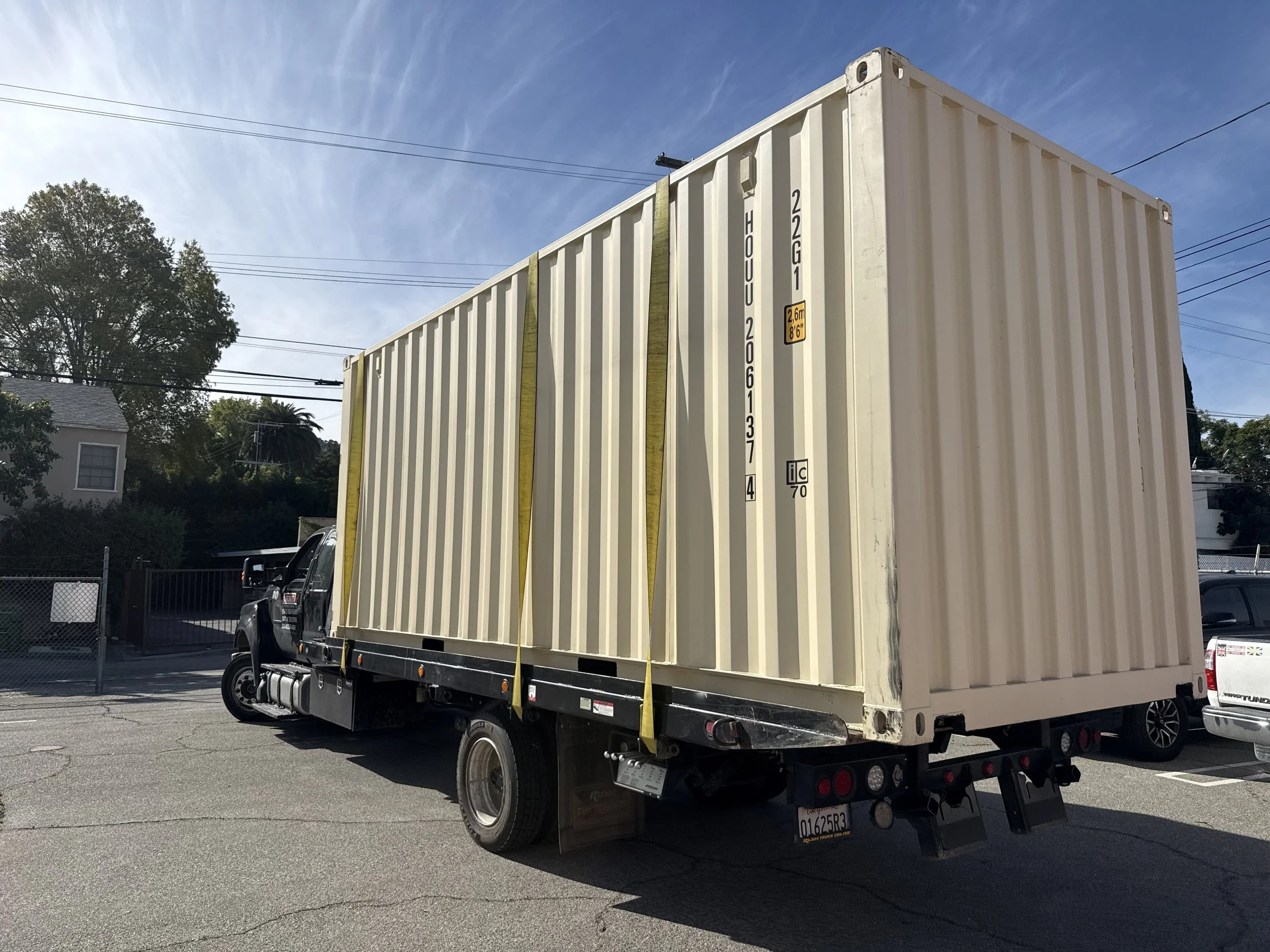 A large delivery truck with a beige shipping container parked on a paved lot, with residential buildings, trees, and power lines in the background under a clear blue sky.