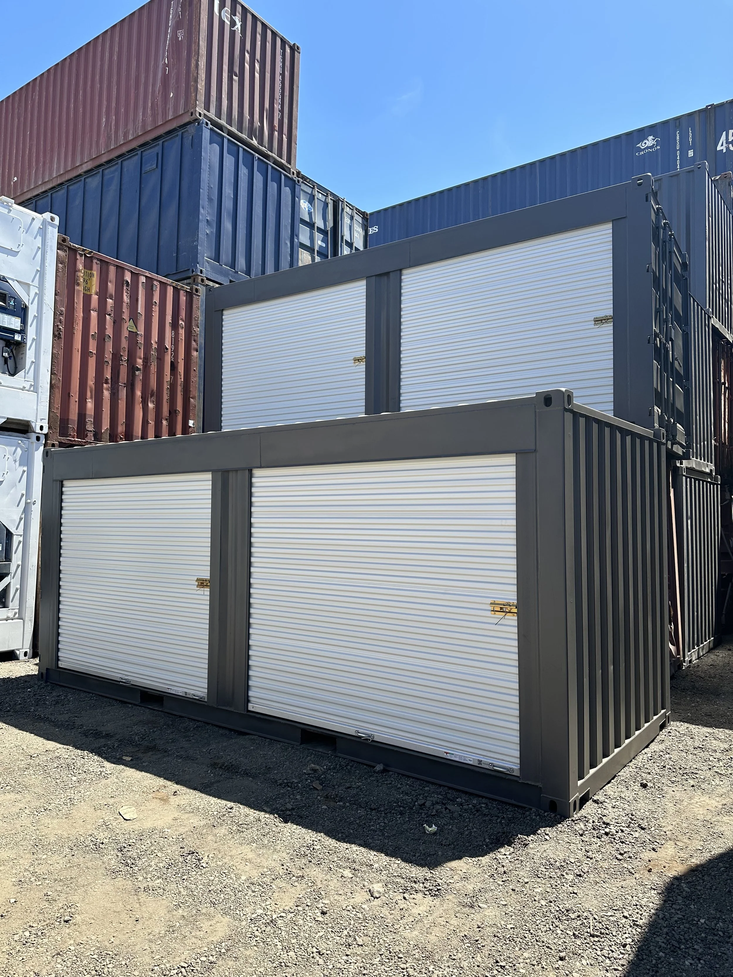 Storage containers stacked outdoors under a clear blue sky, including a white container with rolling doors in the foreground and colored containers in the background.