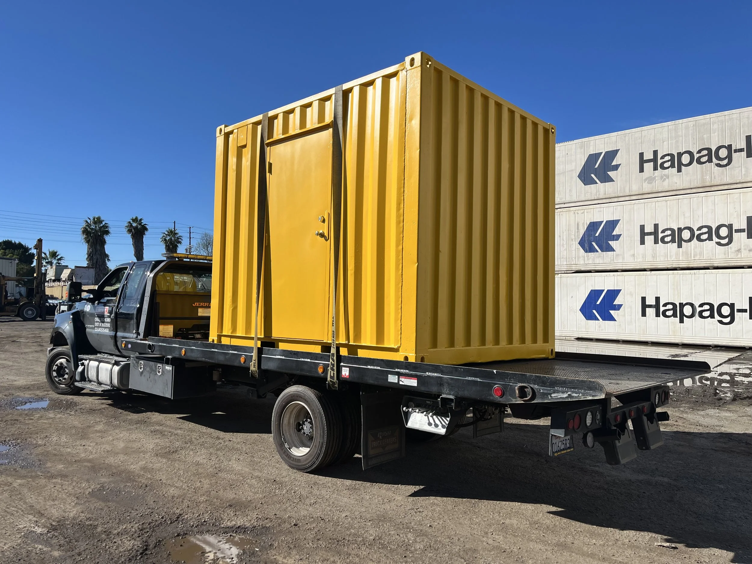 Flatbed truck with a yellow container on the back, parked on a dirt lot with a shipping containers in the background.