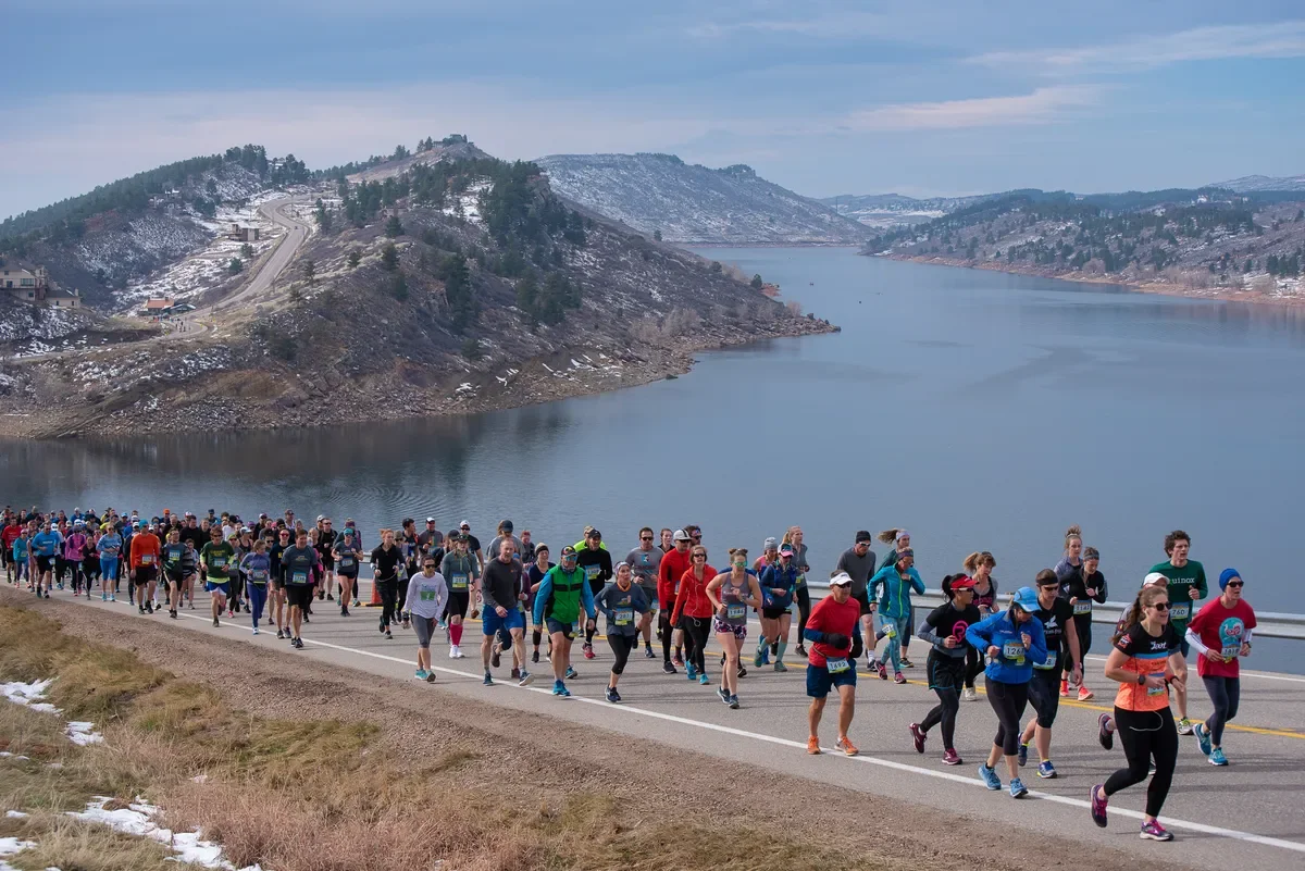 A large group of runners participating in a marathon along a scenic lake with hills and snow patches in the background.