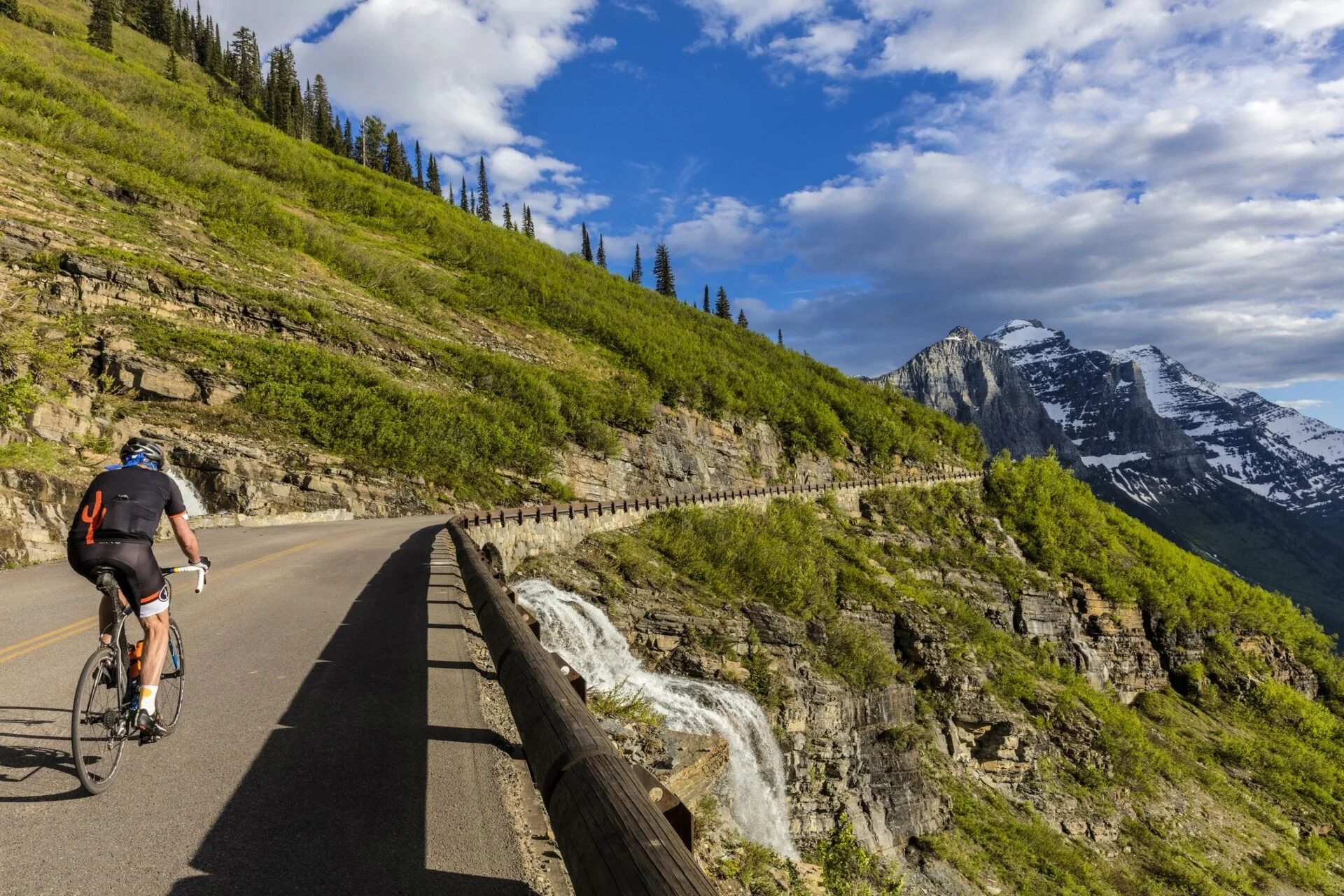 A cyclist riding along a mountain road with a waterfall on the side, green trees on a hillside, snow-capped mountains in the background, and a partly cloudy sky.