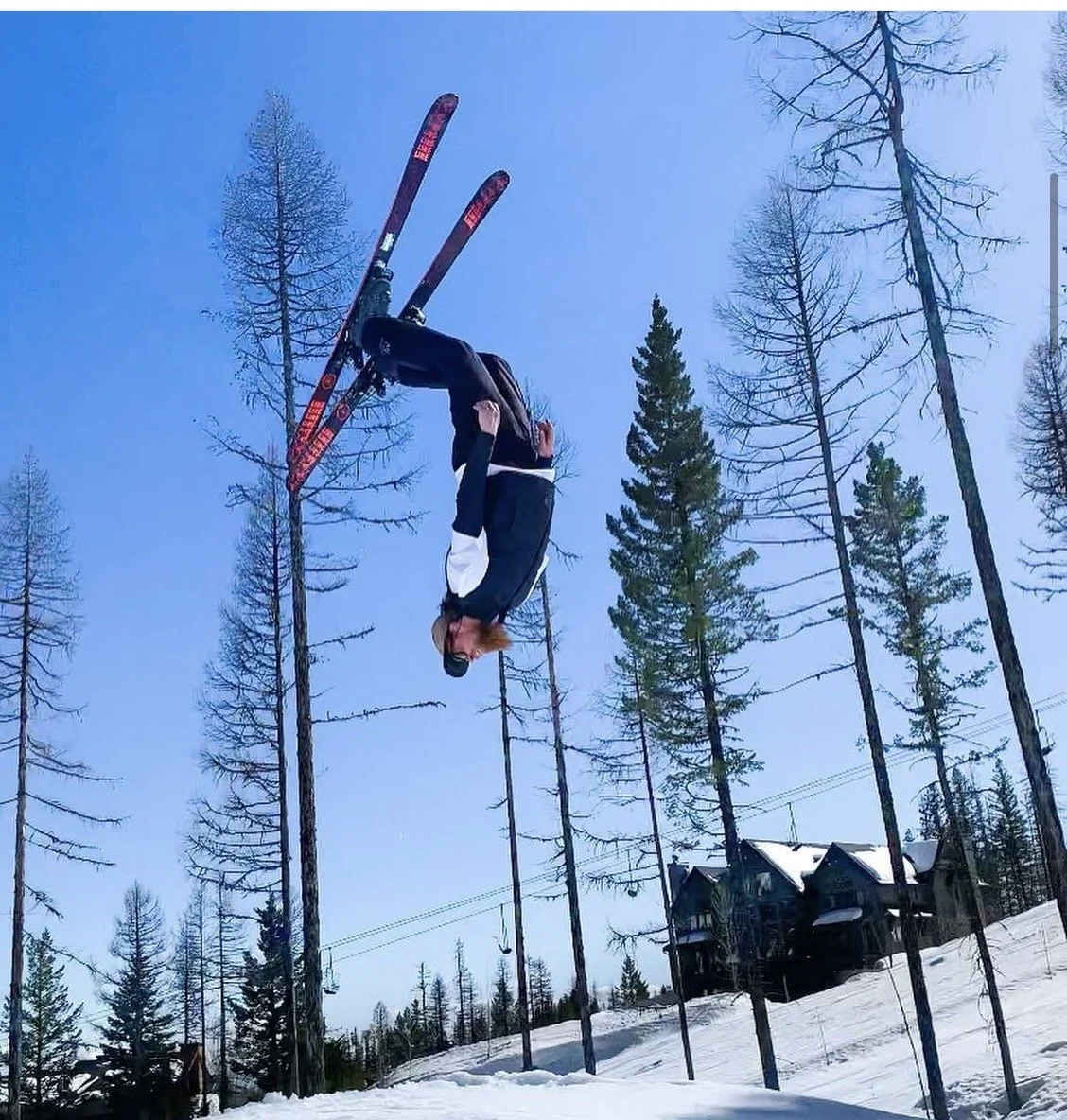A person wearing a black and white jacket and black pants performing a backflip in mid-air over snow-covered ground, with tall, leafless trees and a house in the background under a clear blue sky.