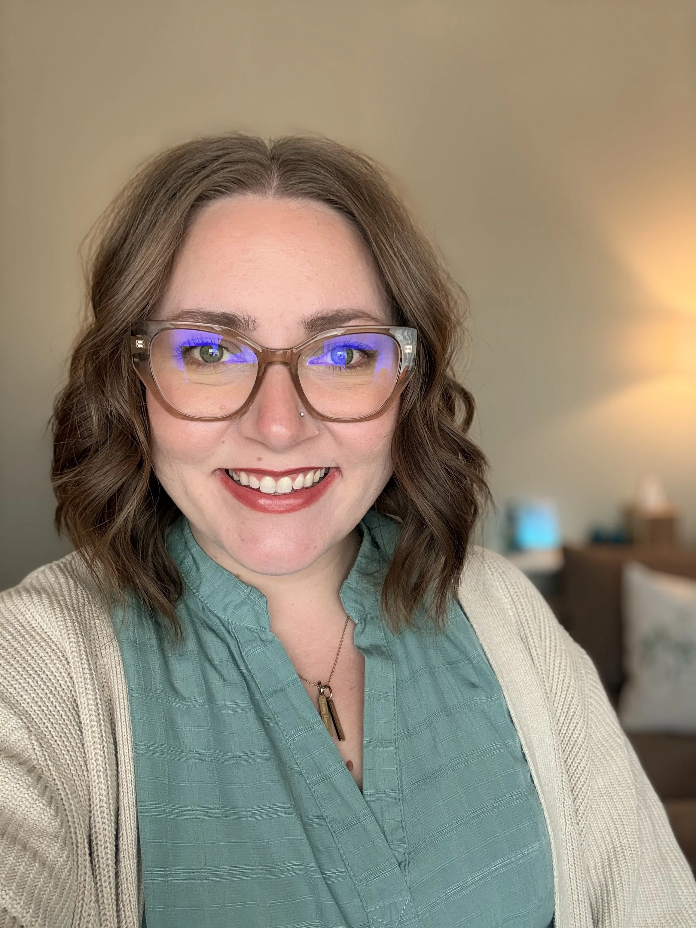 A smiling woman with brown curly hair, glasses, wearing a teal blouse and beige cardigan, indoors with a blurred background.