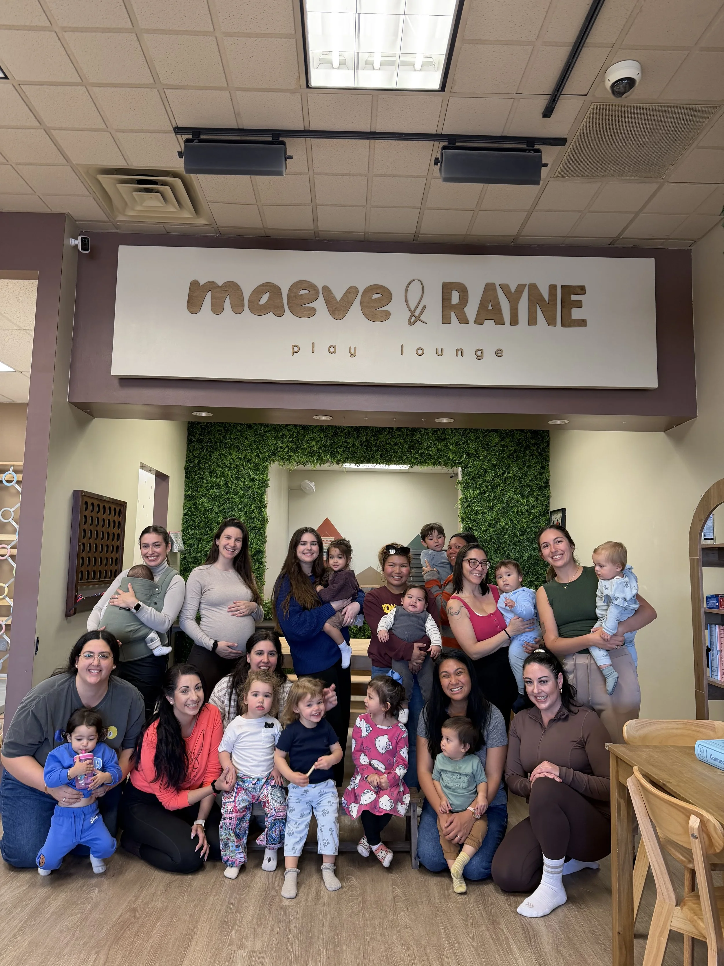 Group of women and children inside a play lounge, posing for a group photo in front of a green wall with the sign 'Maeve & Rayne Play Lounge' above them.